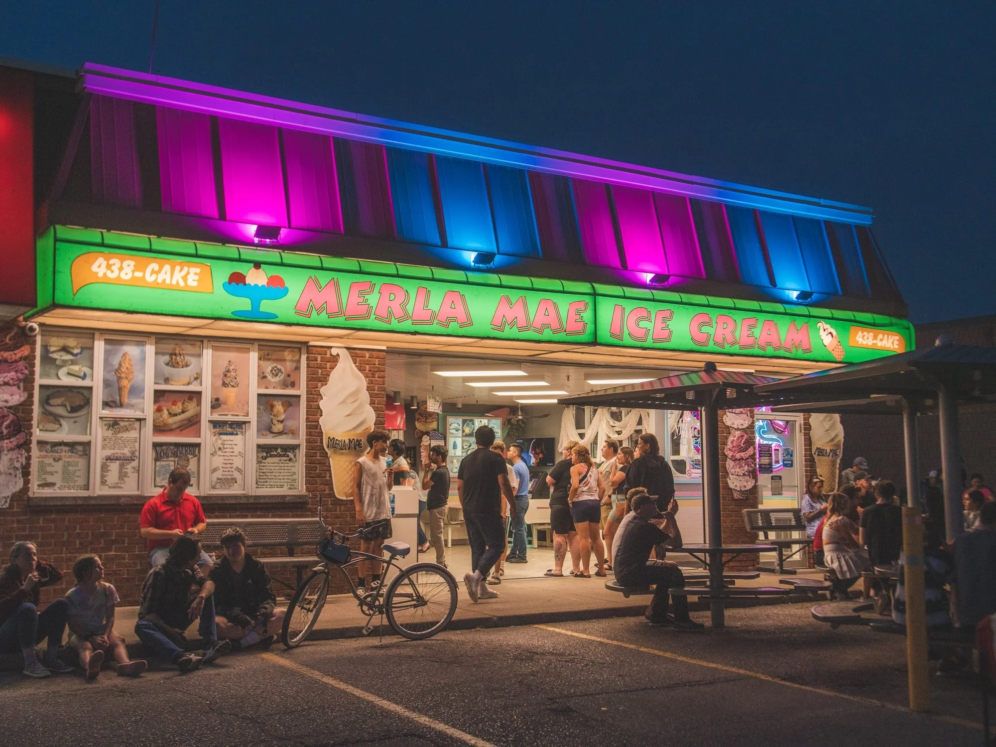 Night scene of Merla Mae Ice Cream shop with neon signage, colorful lighting, and people gathered outside. Some are sitting, and others are standing, with a bicycle parked nearby.