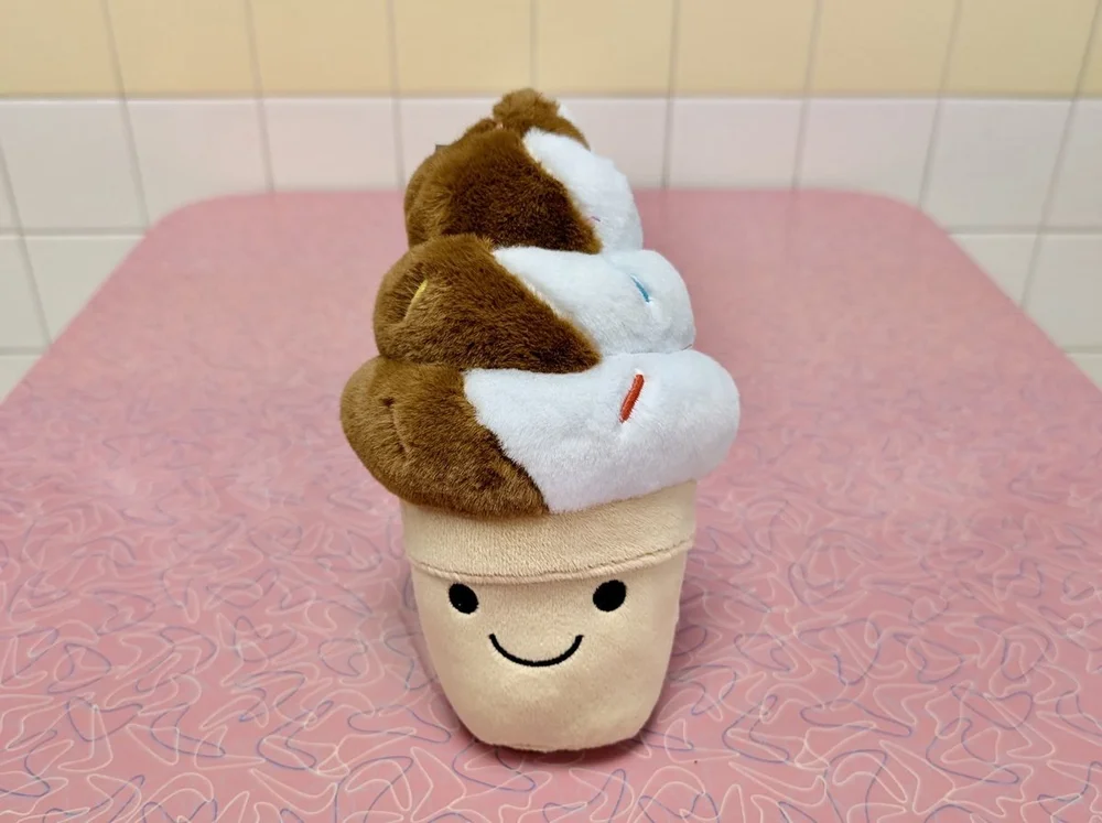 Smiling soft-serve cone plush toy on a pink boomerang-pattern table inside Merla-Mae Ice Cream in London, Ontario