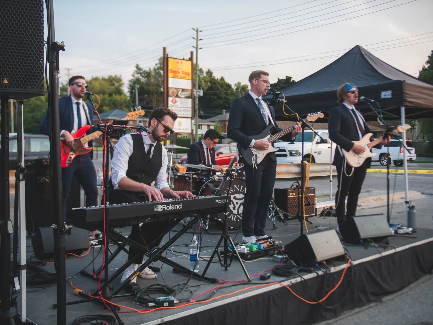Turning Corners in suits playing keys, guitars, bass and drums on the parking-lot stage at National Ice Cream Day 2024, Merla-Mae Ice Cream in London, Ontario