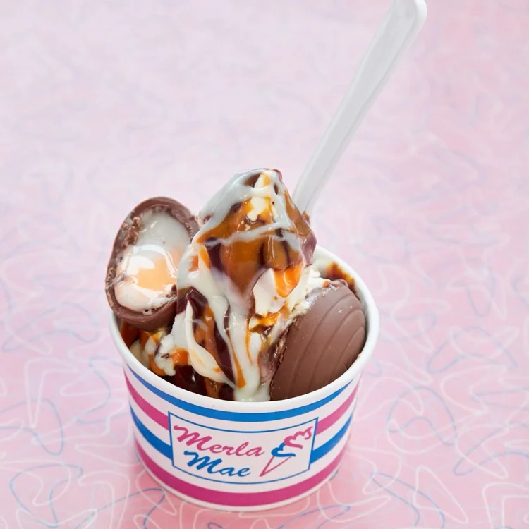 Creme Egg Sundae: vanilla soft-serve drizzled with white, orange and fudge sauces, with the two halves of a cracked Cadbury Creme Egg, in a pink and blue striped cup on a pink boomerang-pattern table at Merla-Mae Ice Cream in London, Ontario