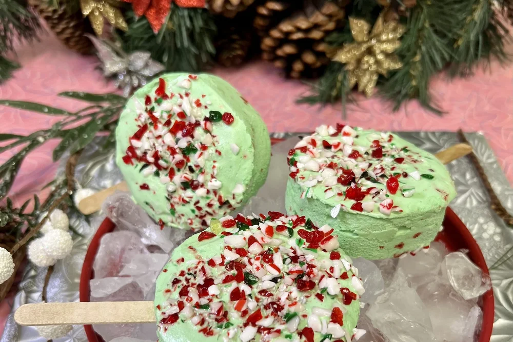 Three mint-green ice cream bars on wooden sticks, topped with crushed red, white and green candy cane, in a red bowl of ice with pine boughs and pinecones, at Merla-Mae Ice Cream in London, Ontario