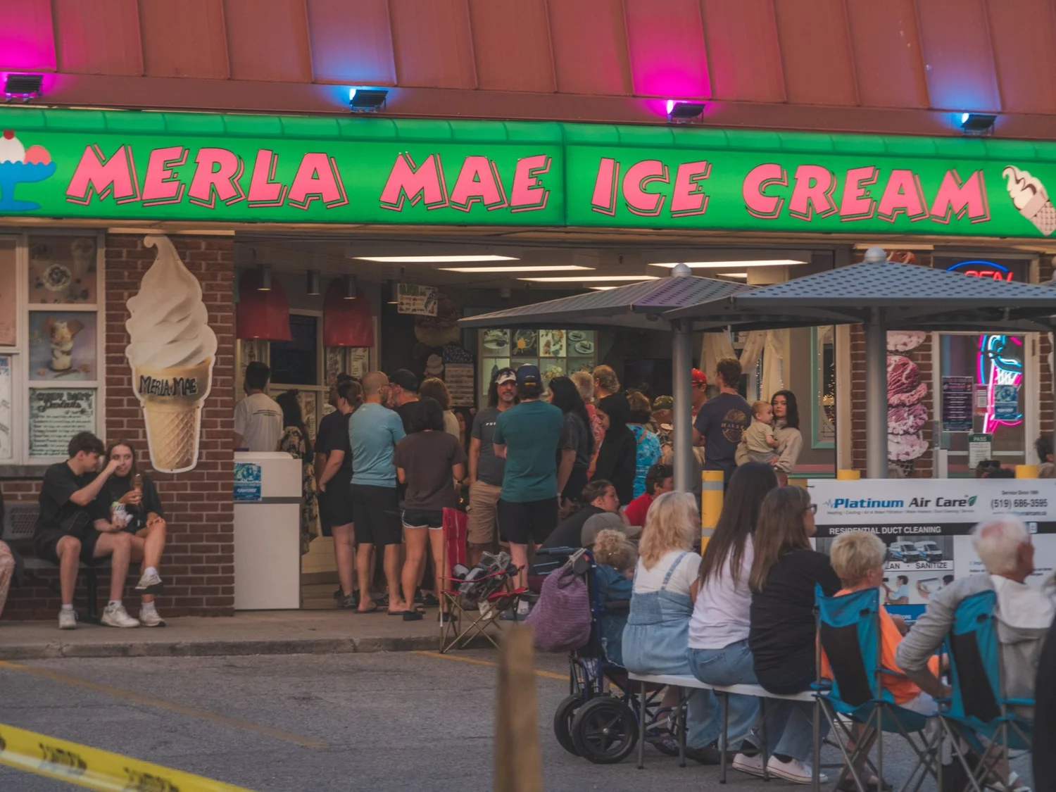 Line out the door at Merla-Mae Ice Cream on National Ice Cream Day, with a local sponsor's sign beside the patio, in London, Ontario
