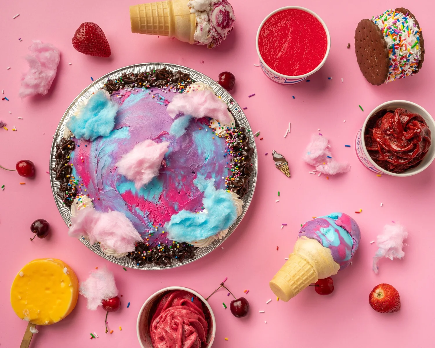 Ice cream party spread on a pink table: an ice cream pie swirled pink, blue and purple and topped with cotton candy, a sprinkle-rolled ice cream sandwich, a dipped ice cream bar on a stick, ice cream cones and cups, at Merla-Mae Ice Cream in London, Ontario