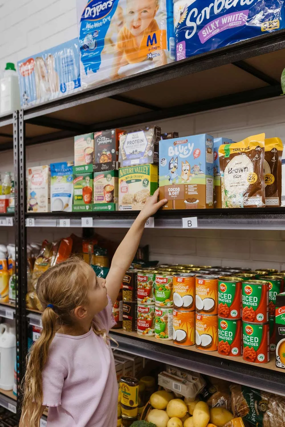 Little girl with long pig tails reaching up to the Bluey cereal box at a Pantry shop