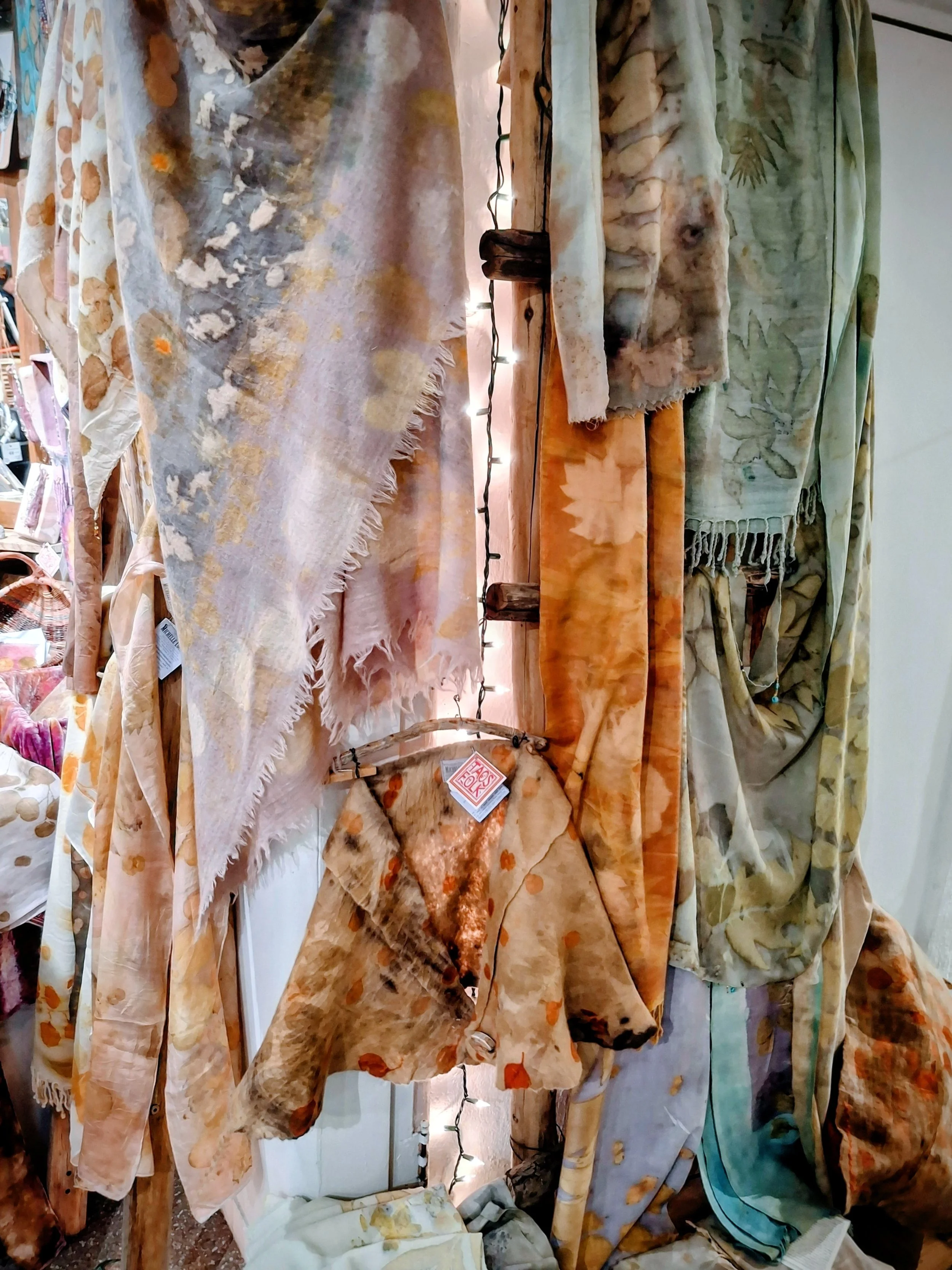 Display of tie-dye and batik fabric scarves hanging on a wooden display with string lights.