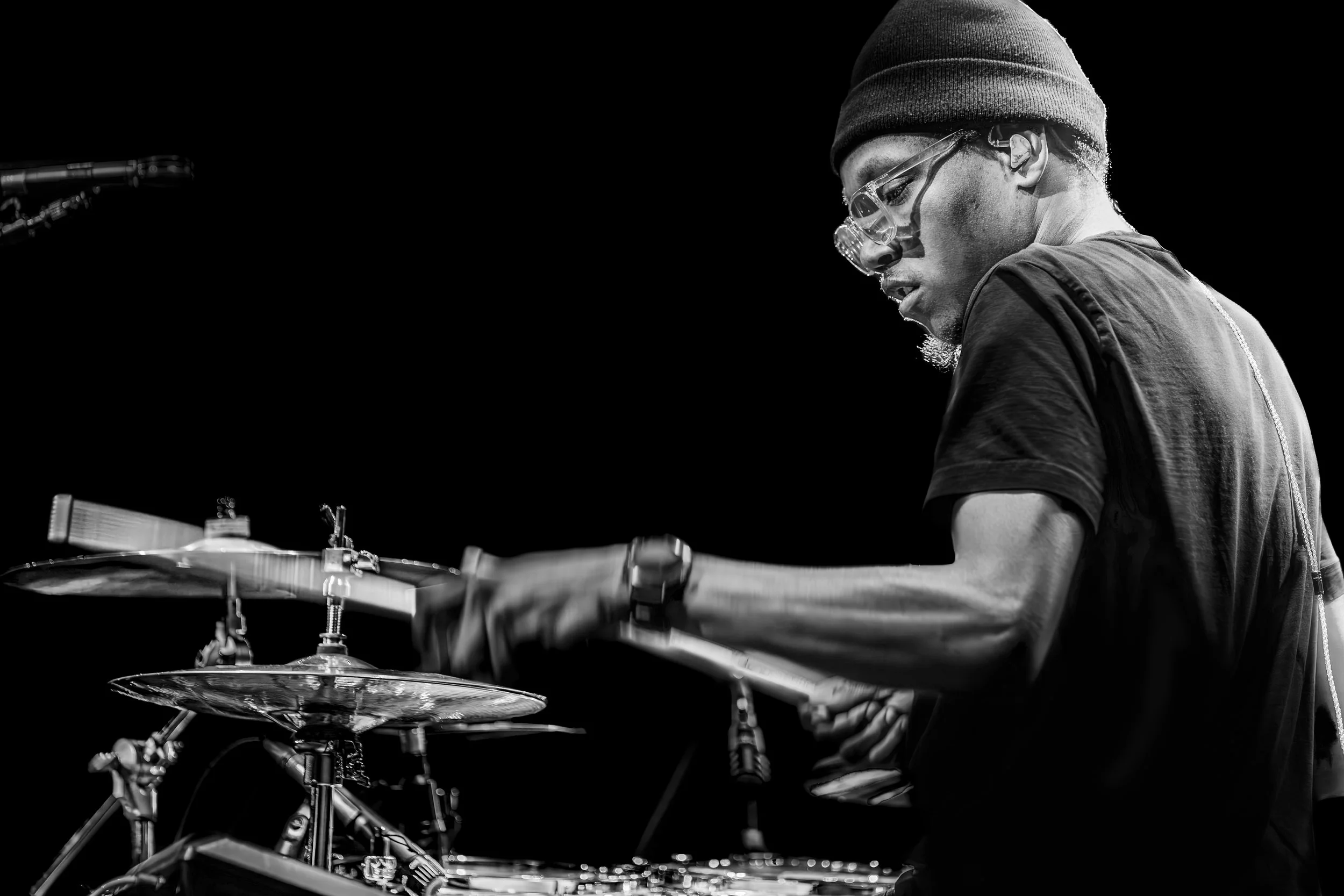 A musician playing the drums, wearing glasses, a beanie, and a T-shirt, captured in black and white.