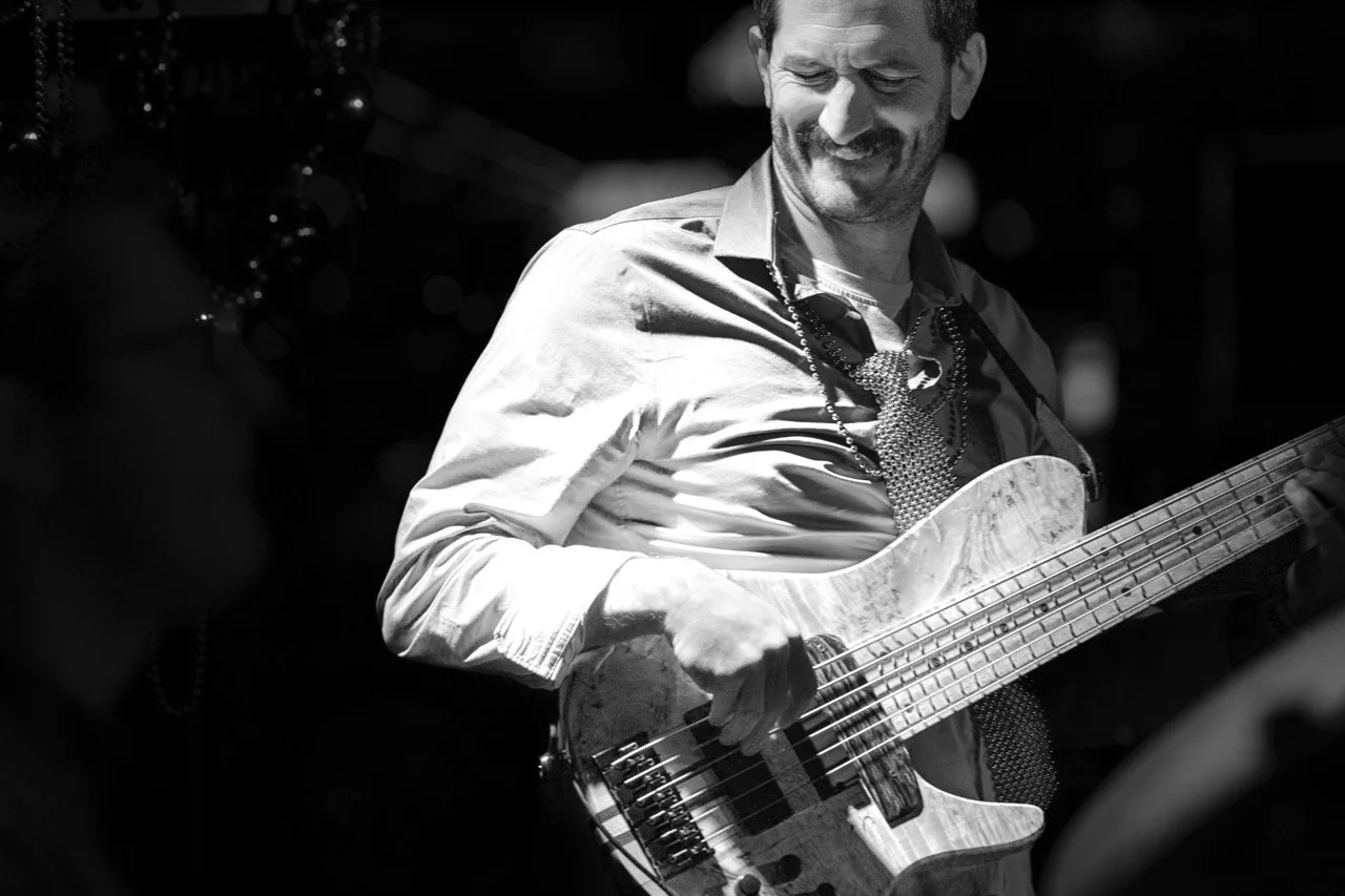 A man playing an acoustic guitar and smiling, wearing a light-colored shirt and necklace, in a setting with a dark background.