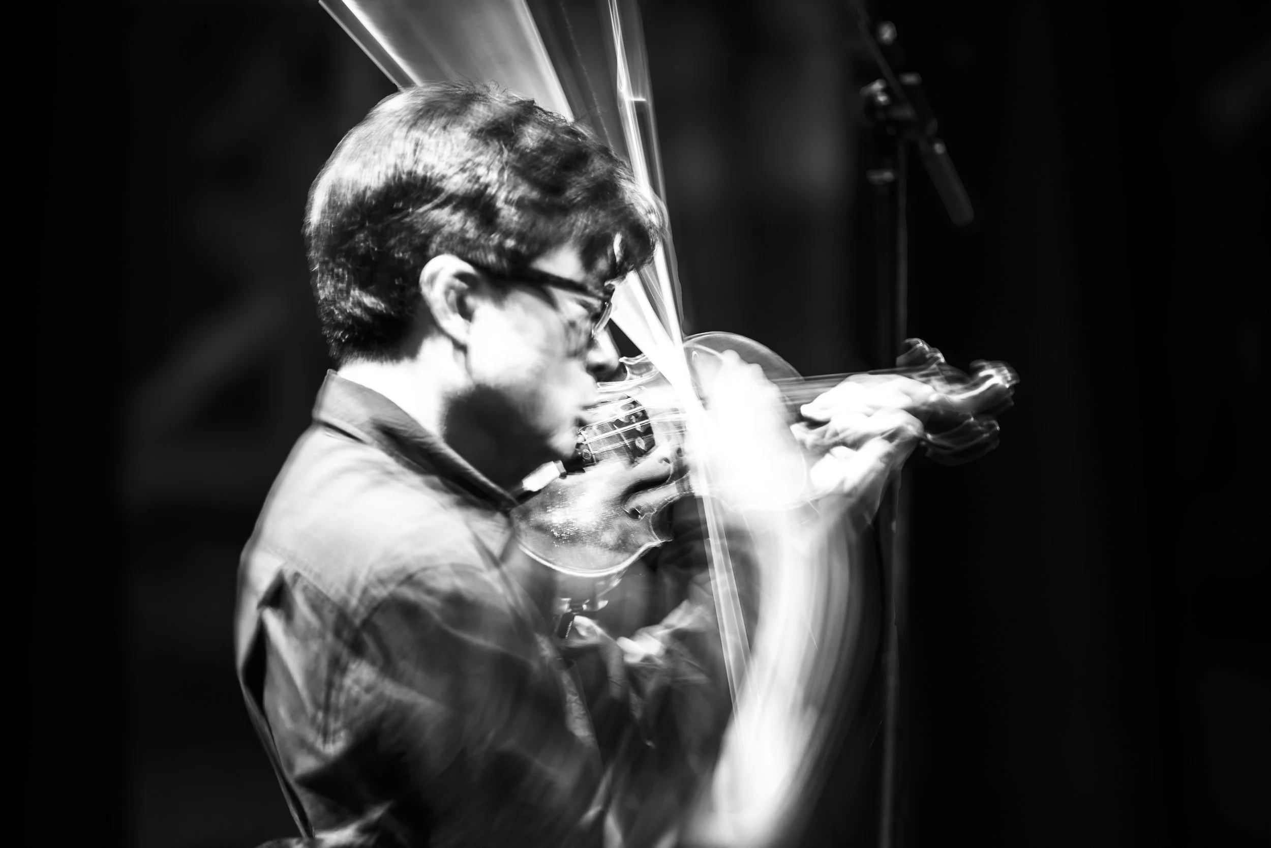 Black and white photo of a man playing the violin with a serious expression, focusing on his face and the violin, against a dark background.