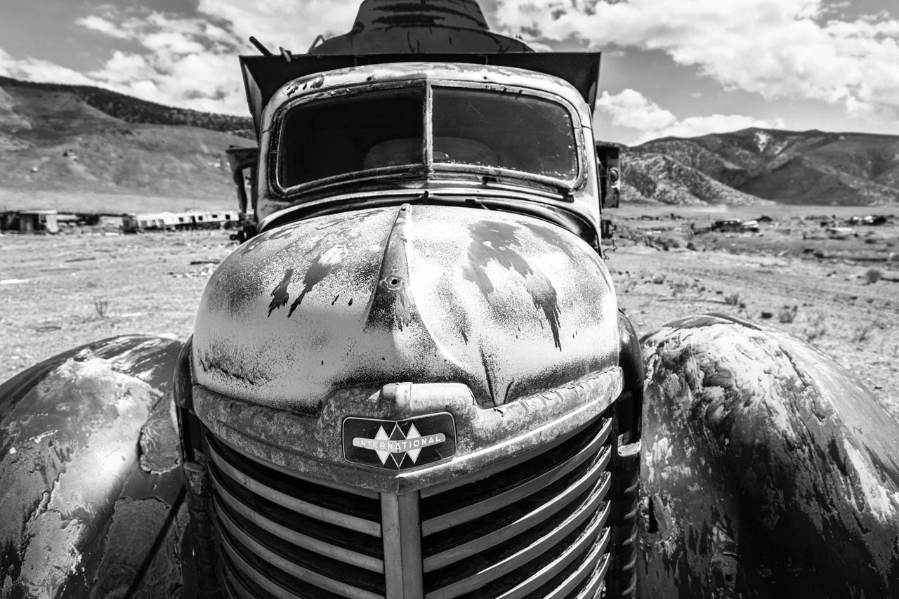 A black and white photo of an old, weathered truck with a rounded front and large fenders. The truck is in a desert landscape with mountains in the background, and the sky has scattered clouds.