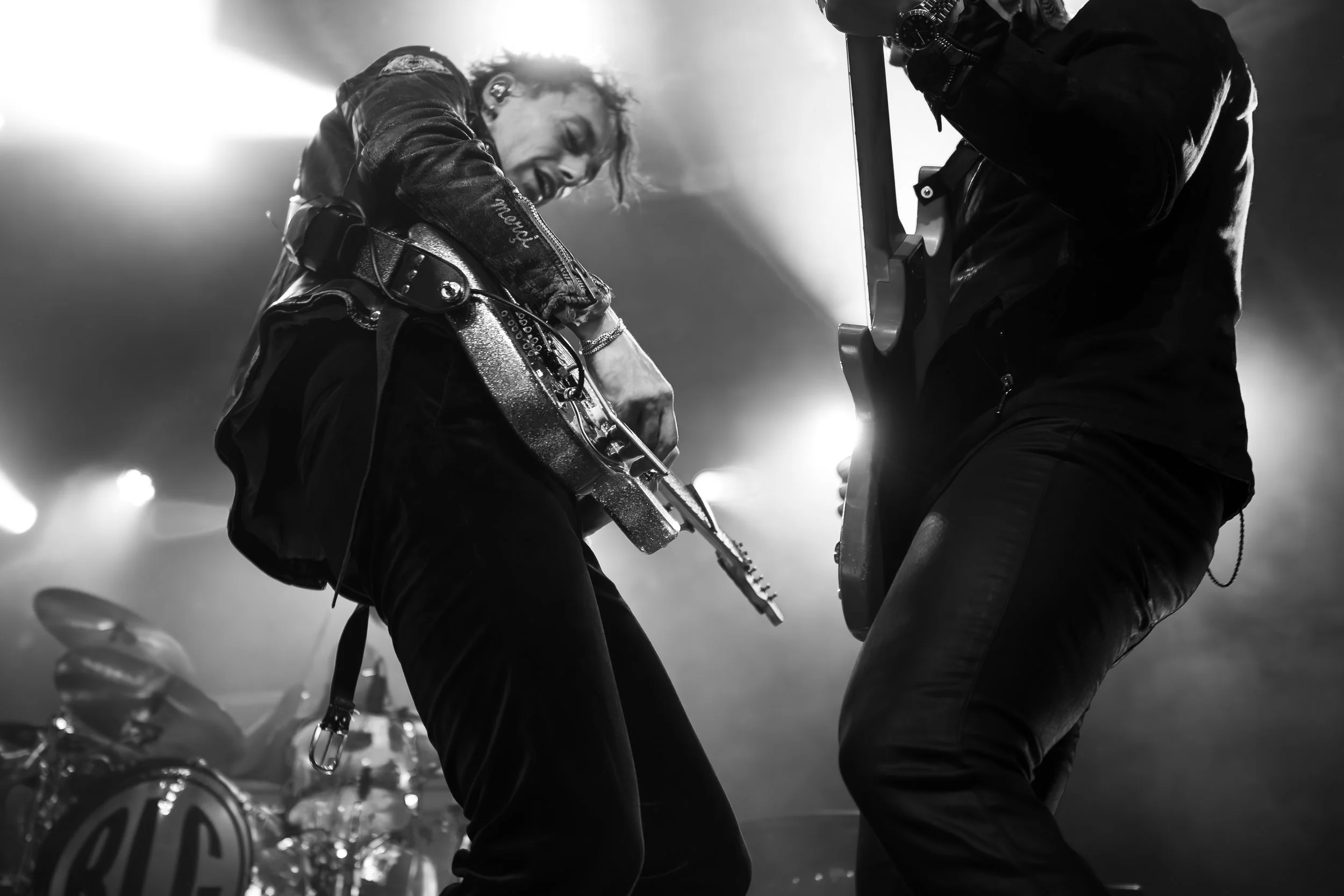 Black and white photo of two people playing electric guitar and bass guitar on stage, with bright lights behind them.