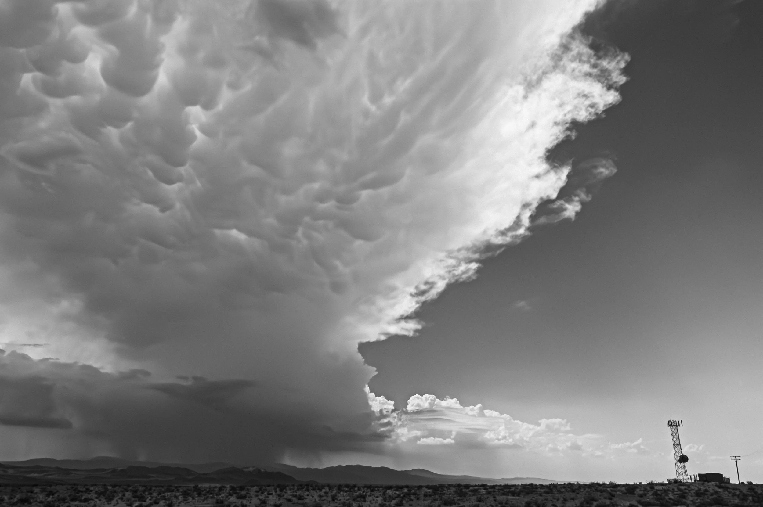 Black and white photograph of a vast open landscape with a dramatic sky featuring large, textured clouds. A communications tower is visible on the right side, and distant mountains or hills stretch across the horizon.
