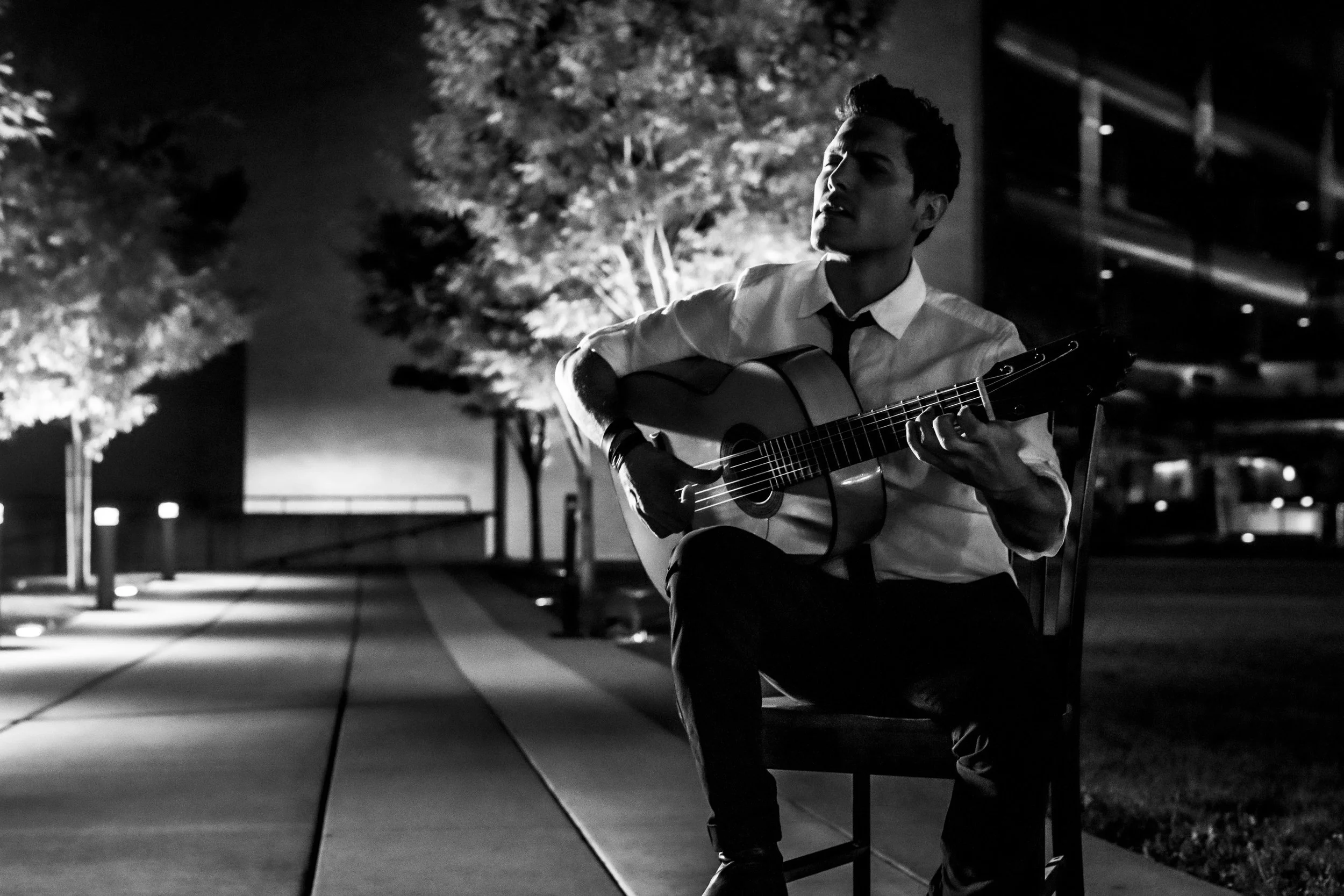 A young man singing and playing guitar outdoors at night, sitting on a chair, with trees and streetlights in the background.