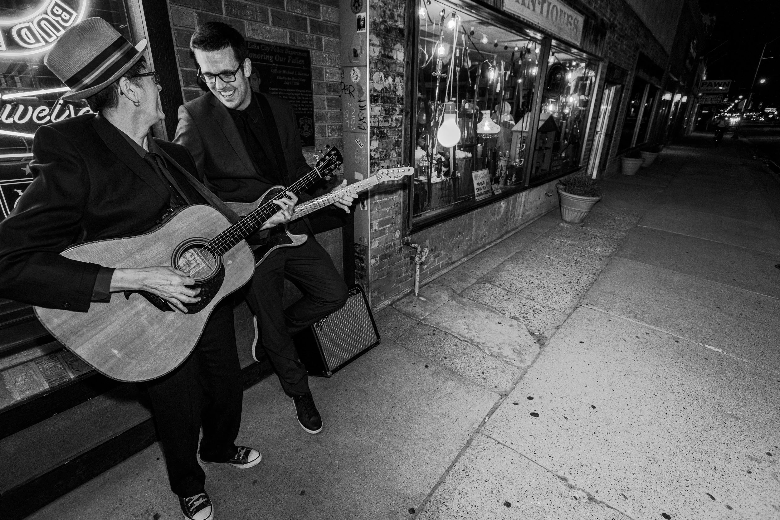Two people are playing guitars on a city sidewalk at night. They are smiling and appear to be enjoying their performance.