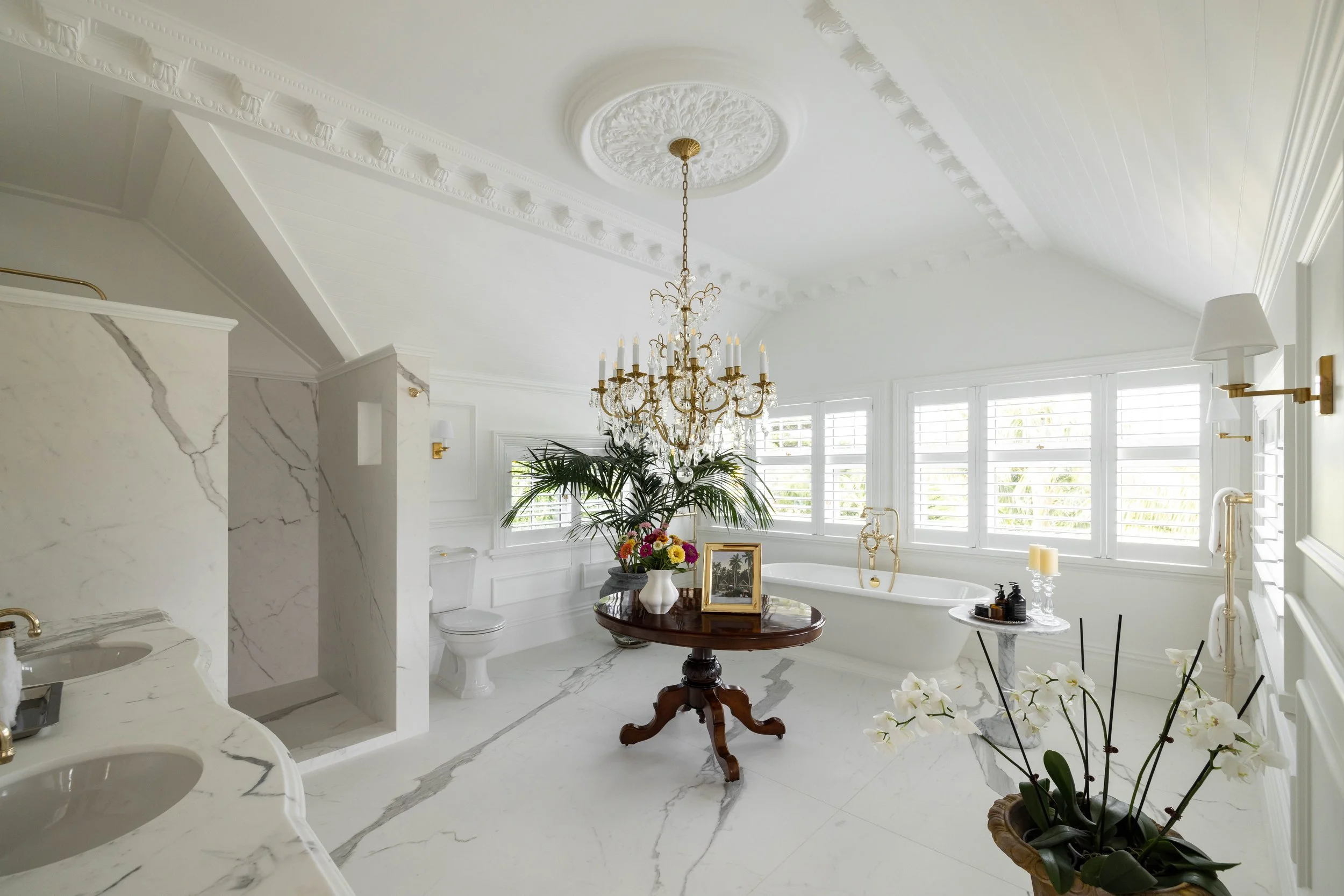 Elegant white bathroom with a chandelier, bathtub, and multiple windows with shutters, decorated with plants, candles, and framed pictures.