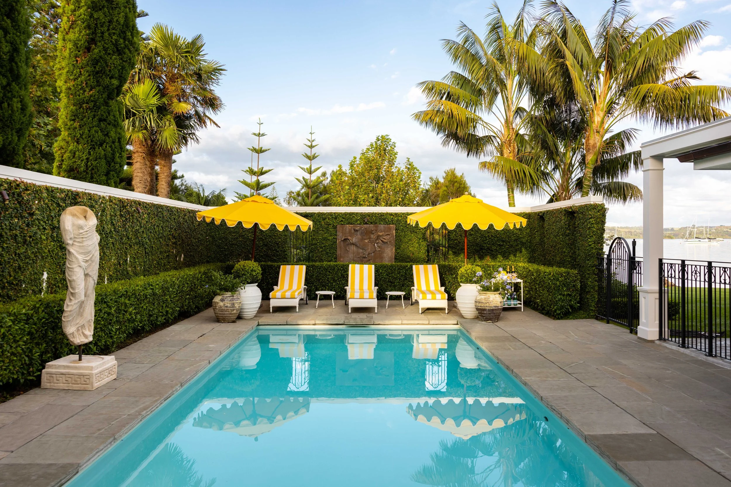 A backyard swimming pool area with two yellow umbrellas, four chairs with yellow and white striped cushions, potted plants, stone statues, lush green hedges, palm trees, and a view of the river or lake with sailboats in the distance.