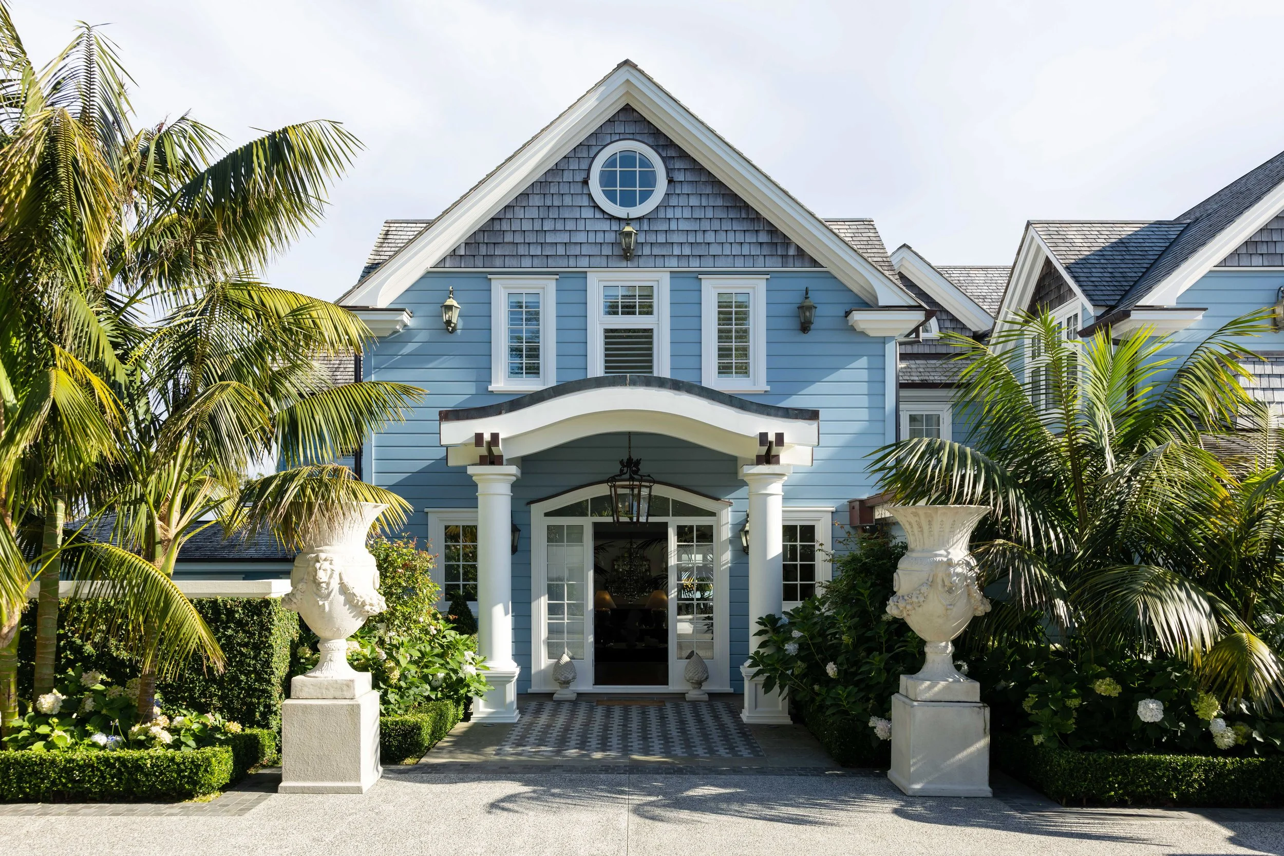 Front view of a blue house with white trim, surrounded by tropical plants and large decorative urns.