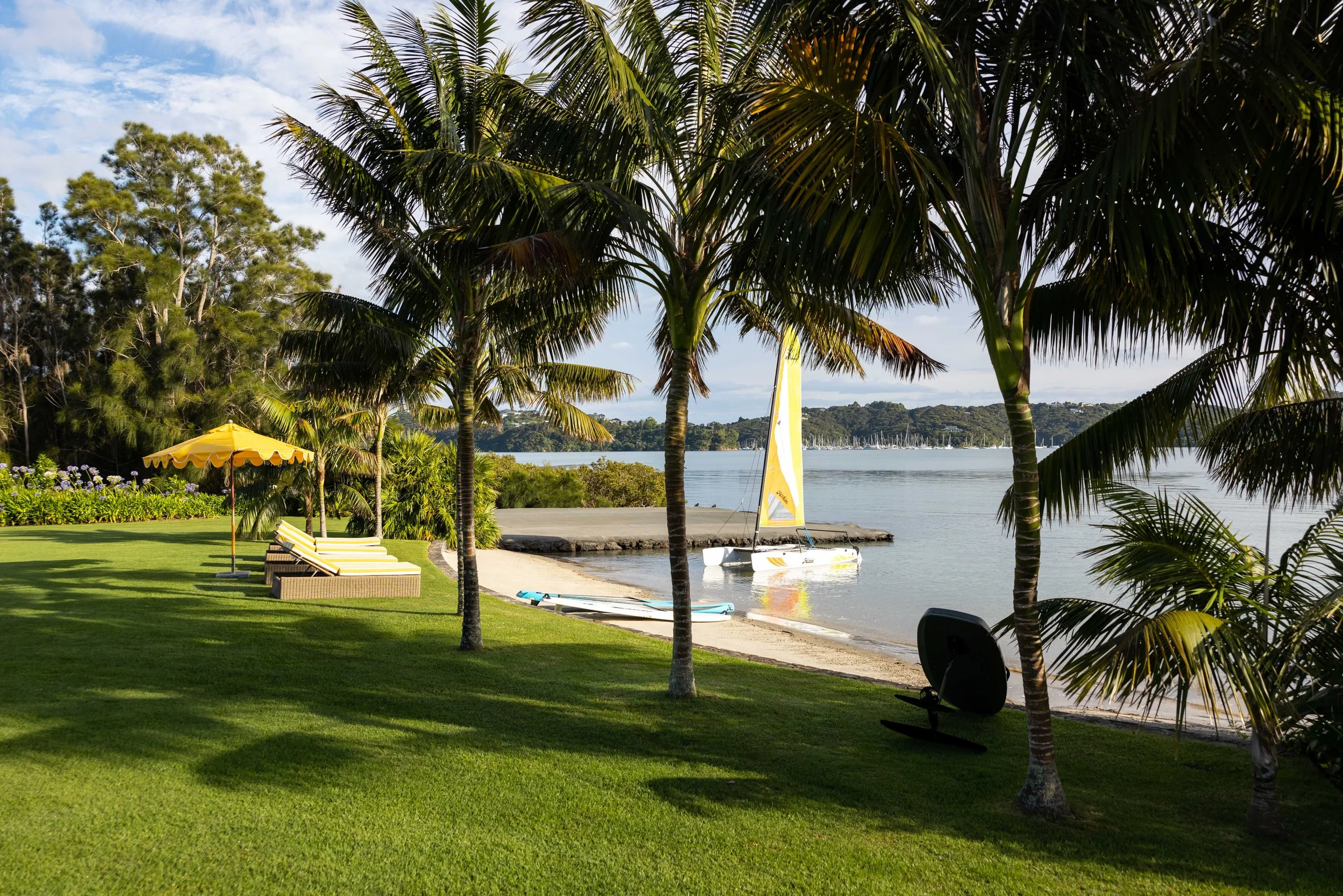 A tropical seaside scene with palm trees, green grass, and lounge chairs under a yellow umbrella. There is a small sailboat and paddleboards by the shoreline, and a calm body of water with a wooded hill in the background.