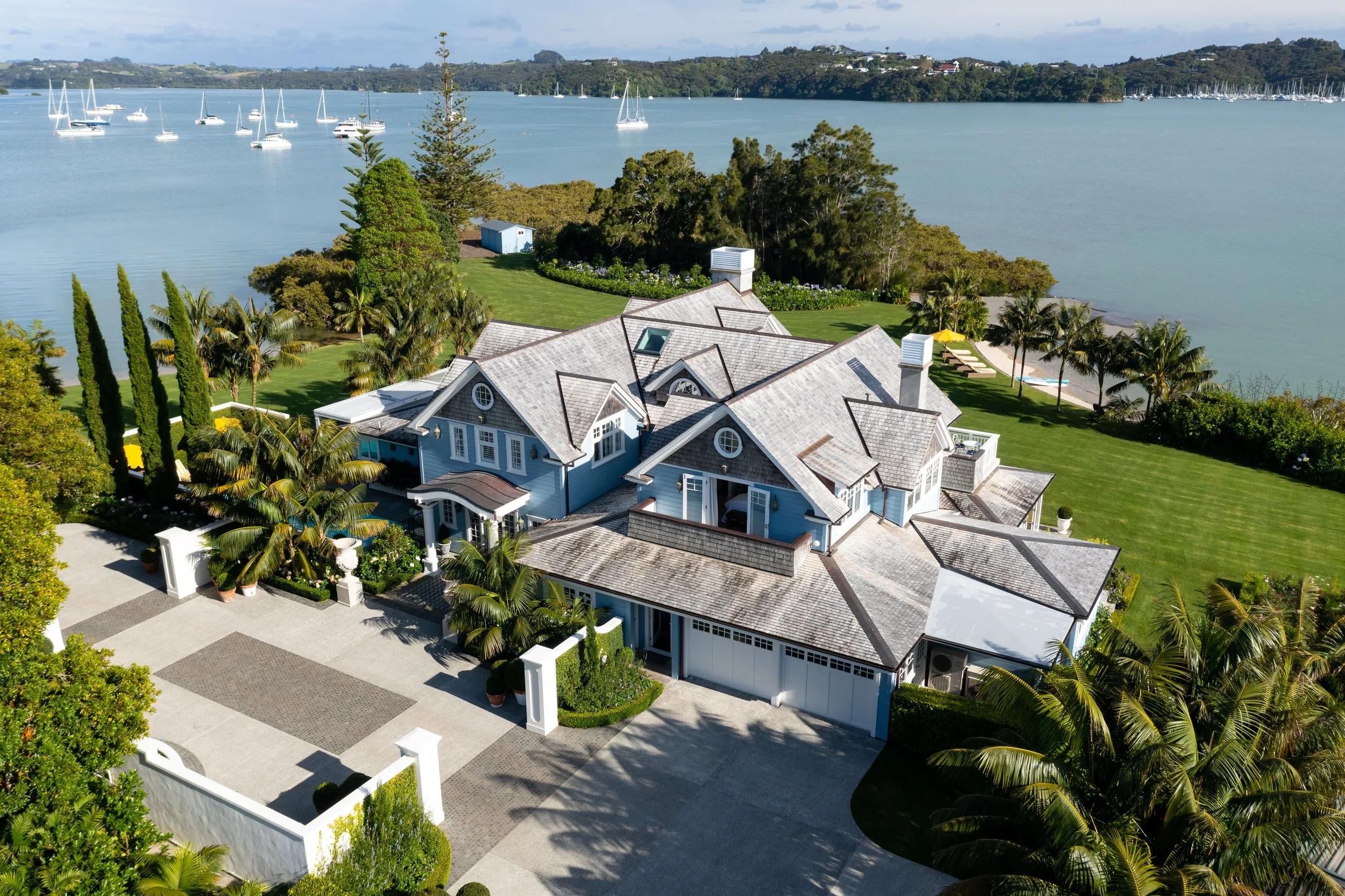 Aerial view of a large waterfront house with a gray shingle roof, surrounded by lush greenery and palm trees, overlooking a body of water with sailboats anchored in the distance.