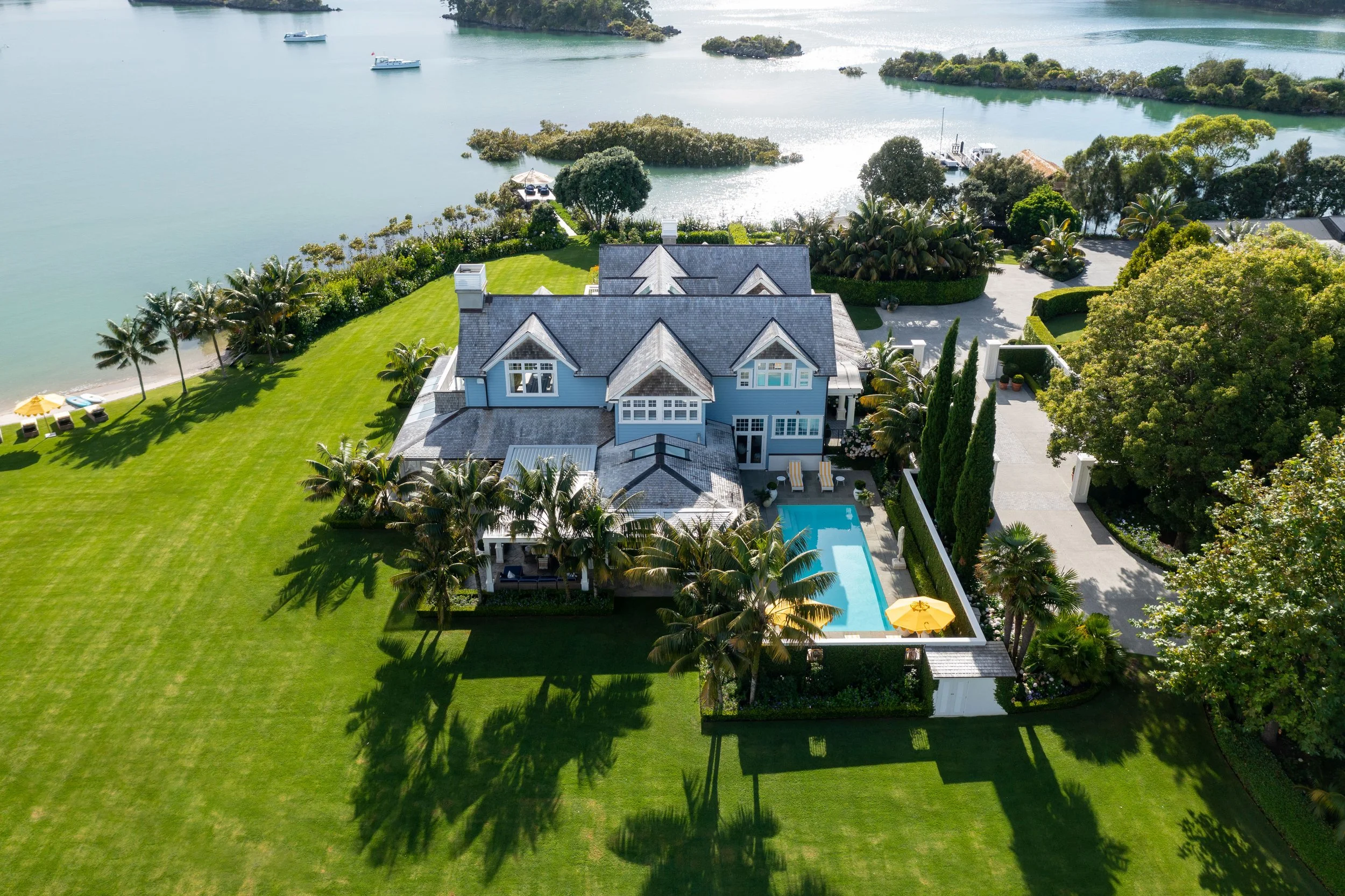 A large, blue, multi-story house with a backyard swimming pool, surrounded by tall palm trees and a green lawn, with a water body and boats in the background.