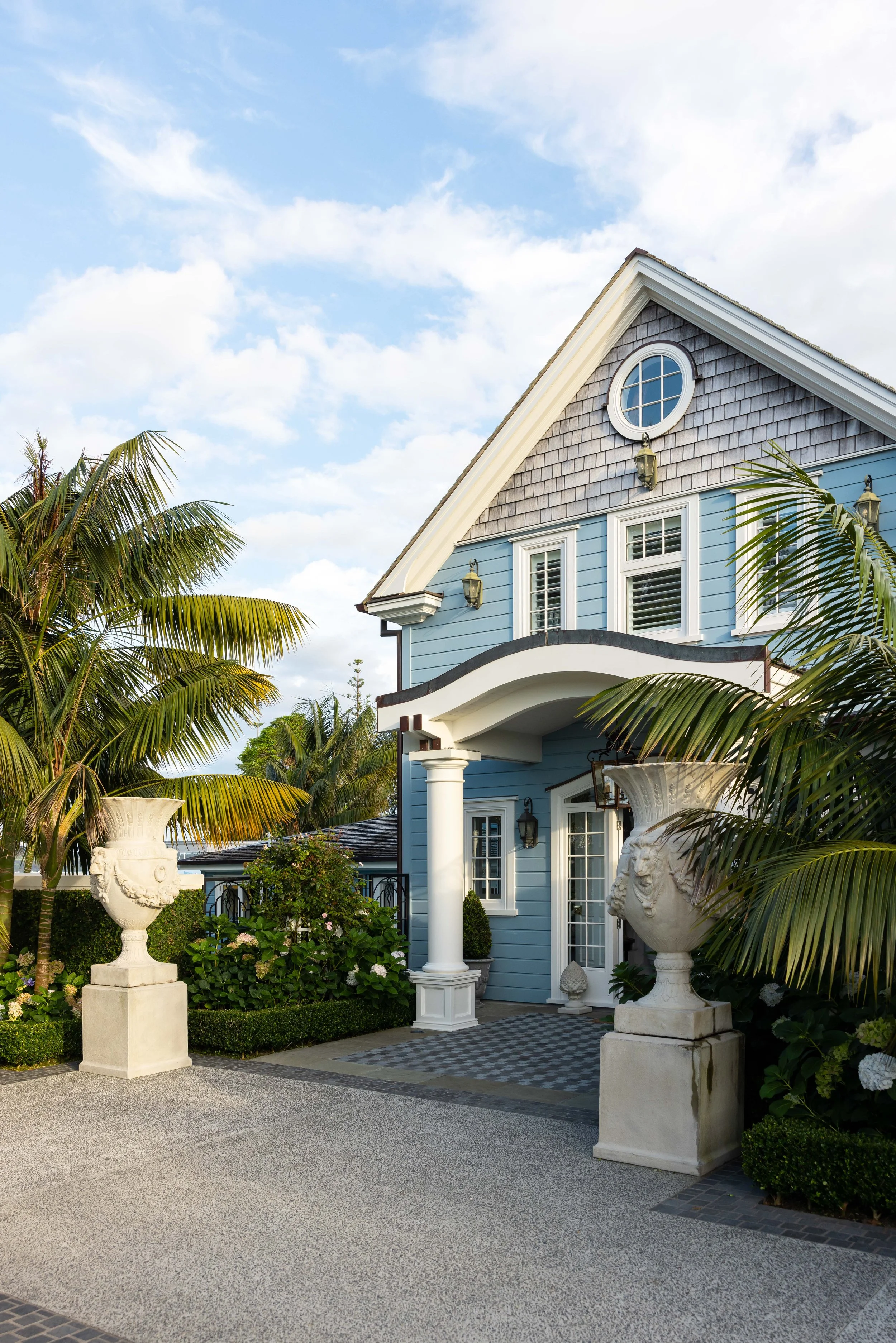 A blue house with white trim, an arched entrance supported by a white column, decorative urns, tropical plants, and a cloudy sky.