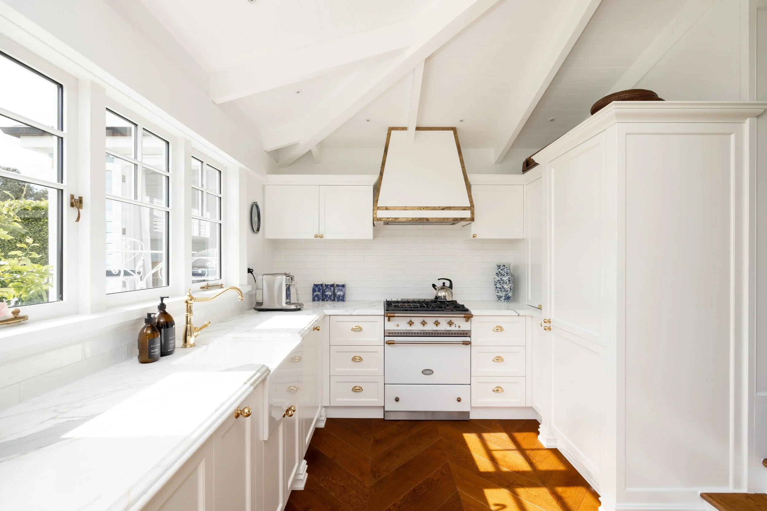 Bright white kitchen with large windows, white cabinetry with brass handles, a marble countertop, and a vintage-style stove with a white range hood, hardwood floor, and decorative vases.