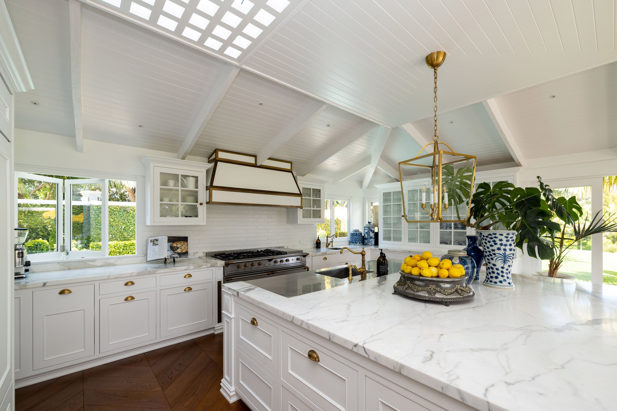 Bright kitchen interior with white cabinets, marble countertops, a large kitchen island, brass rangehood, a brass chandelier, green plants, and windows showing outdoor greenery.