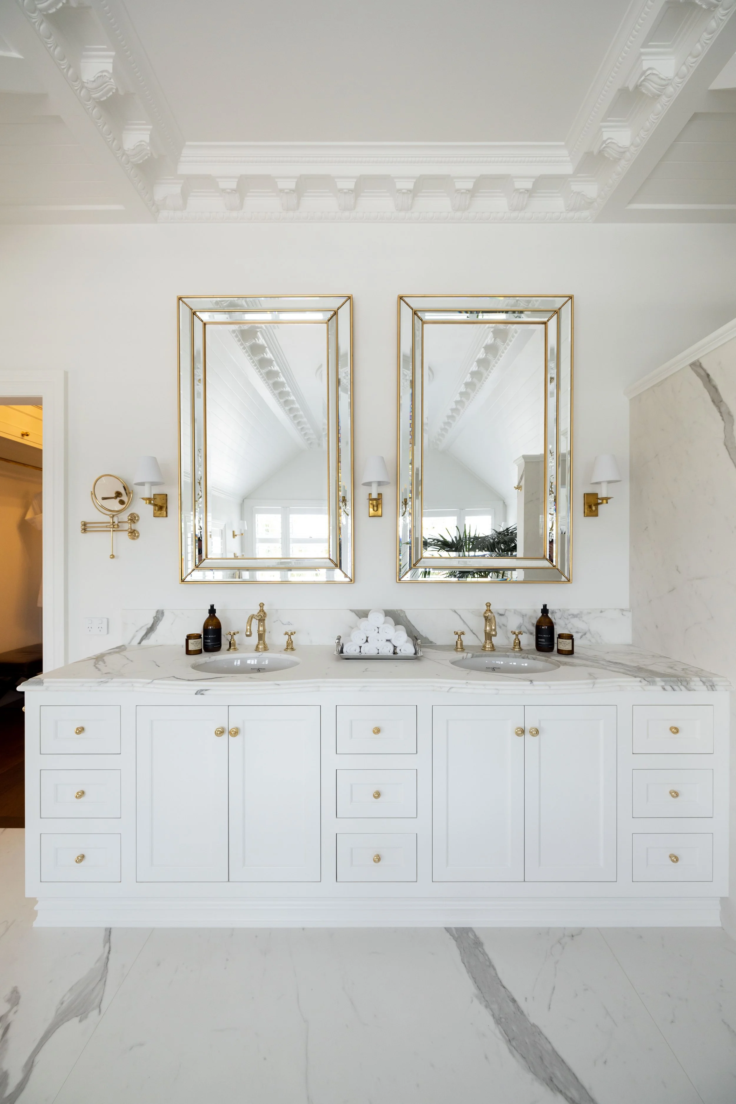 White bathroom vanity with marble countertop, two sinks, and two brass mirrors above, with wall-mounted brass light fixtures, and decor bottles and rolled towels.