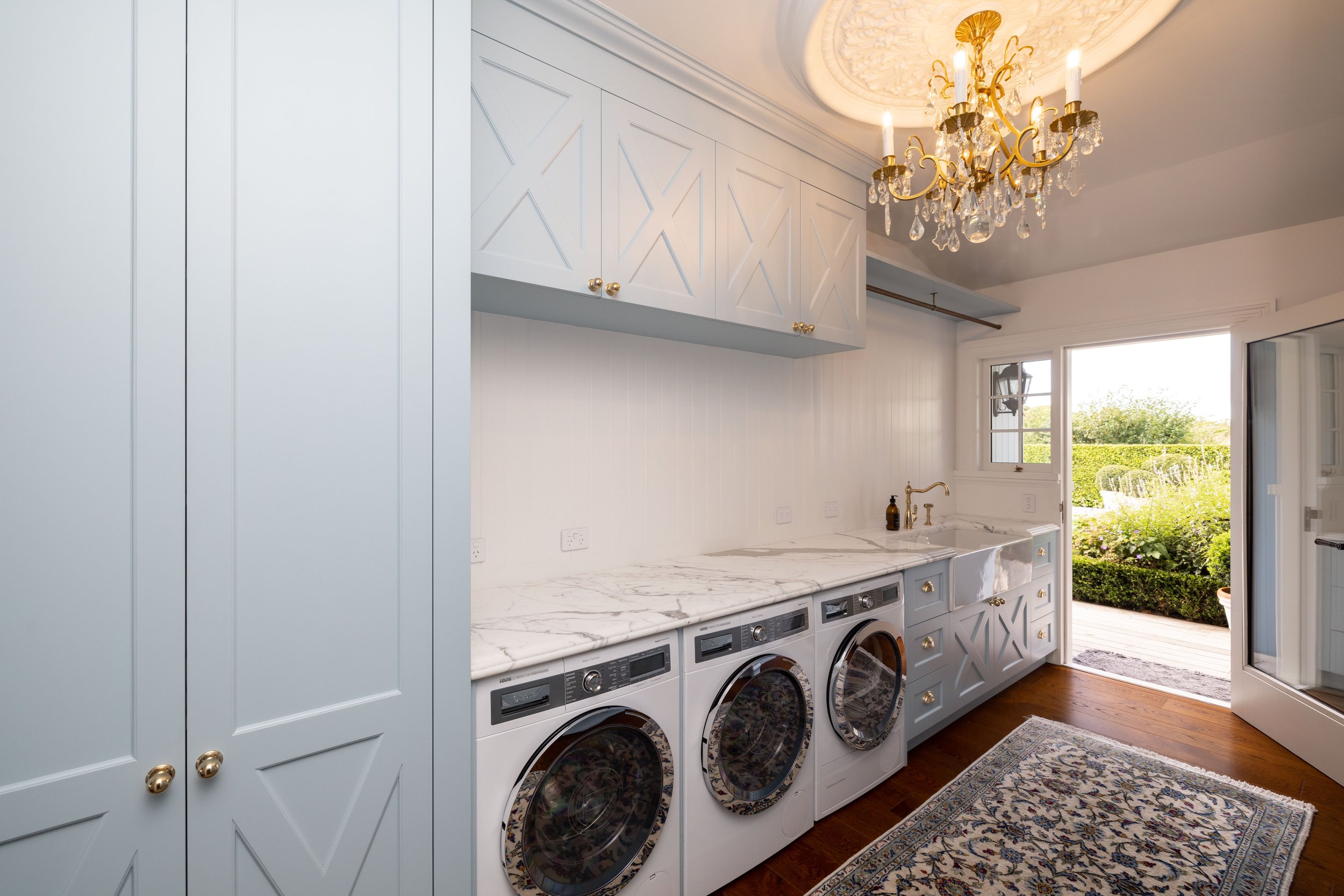 Laundry room with light blue cabinets, marble countertop, front-loading washer and dryer, chandelier, window with outdoor view, and a decorative rug.