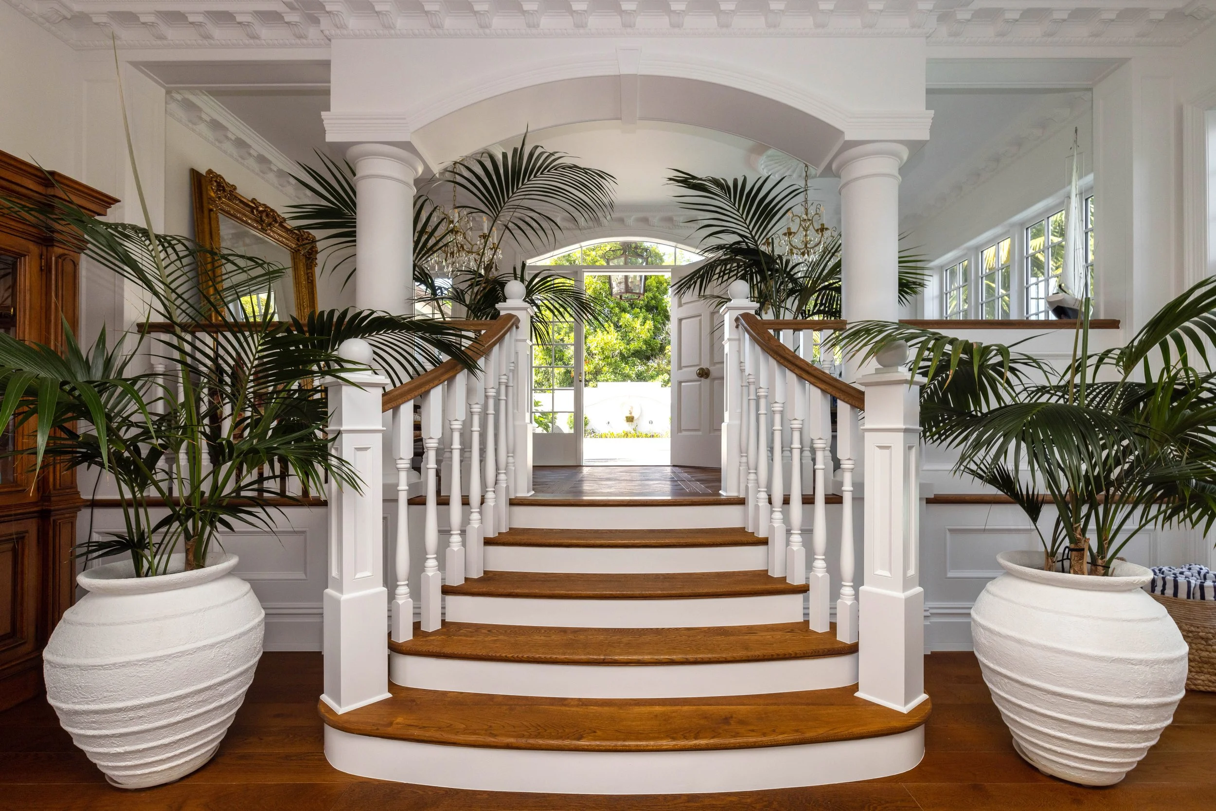 Interior view of a home entrance with white stairs and curved wooden steps, flanked by large potted plants, white walls, ornate molding, and a door with glass panels at the top of the stairs.