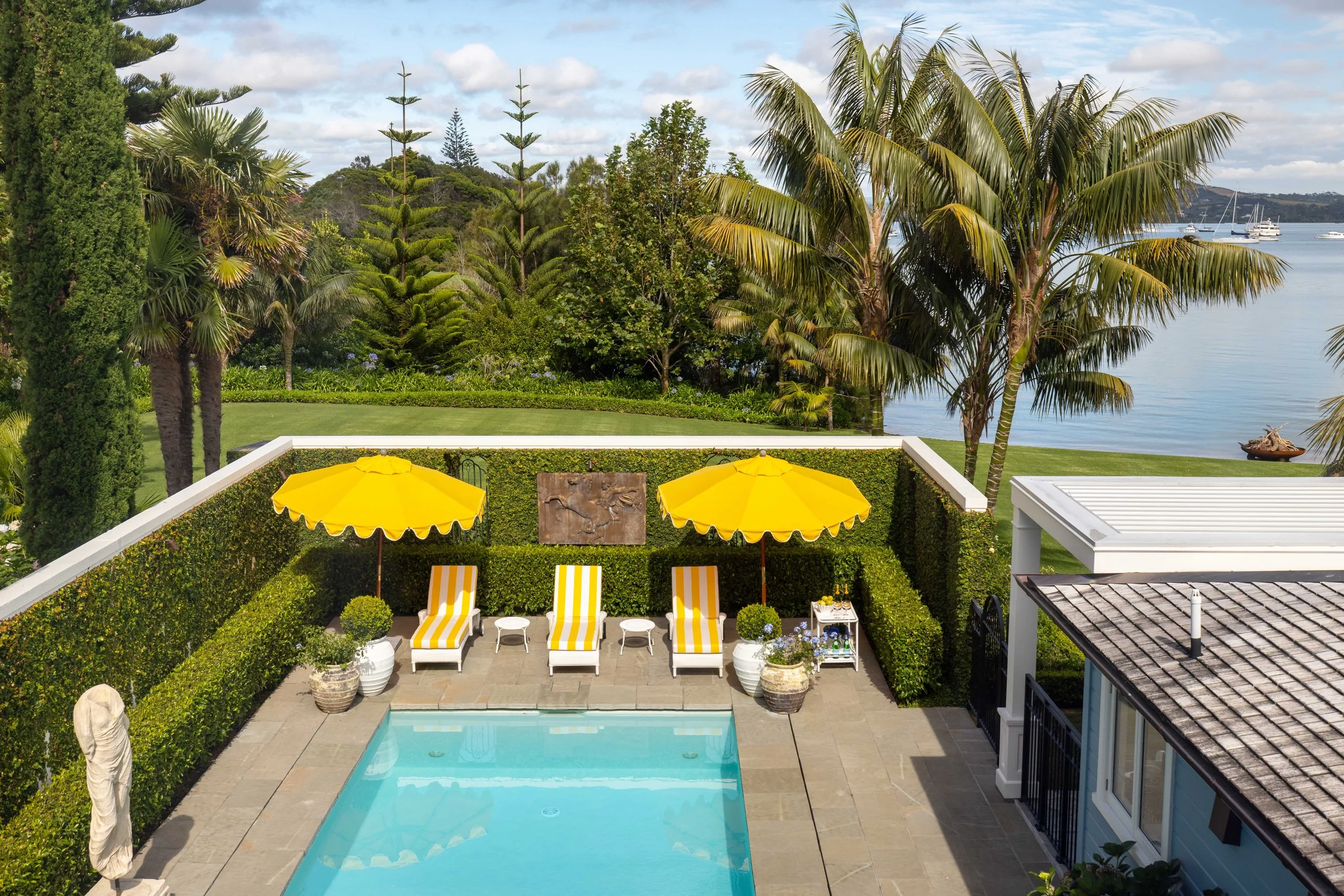 Backyard with a swimming pool, yellow umbrellas, striped lounge chairs, and lush green landscaping near a body of water with boats.