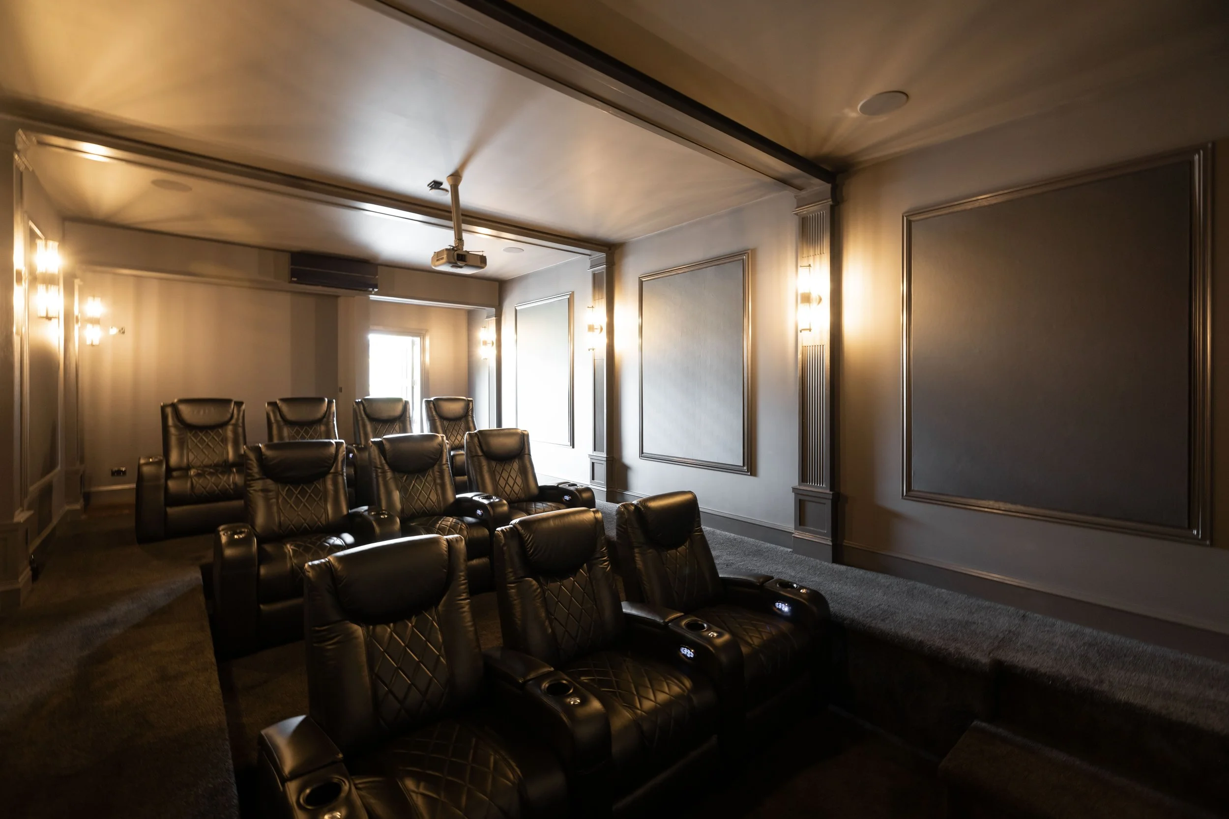 A private home theater room with black leather recliner chairs arranged in two rows, wall-mounted lights, gray walls with decorative molding, and a ceiling-mounted projector.