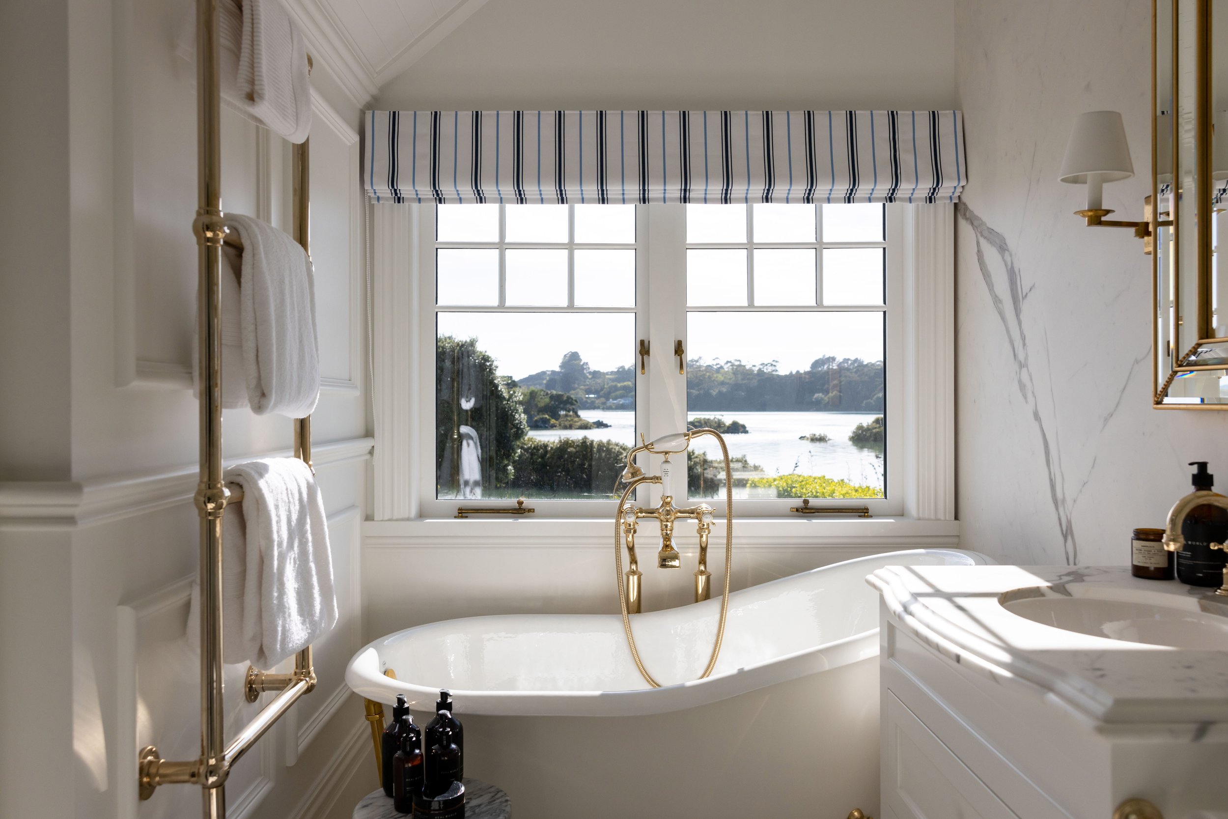 Elegant bathroom with a white bathtub, brass fixtures, a large window with a blue-striped blind showing a river and trees outside, a white marble countertop, and brass towel racks with white towels.