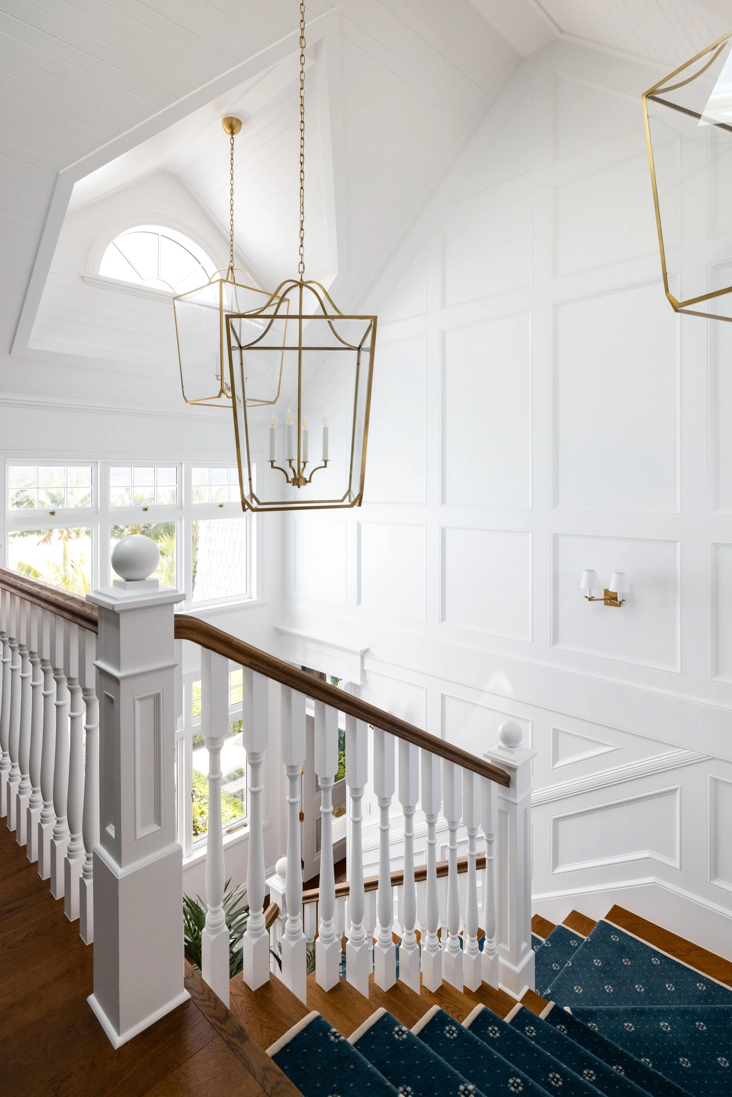 Interior view of a stairway landing with white walls, a wooden handrail, blue carpeted stairs, and modern gold-framed lantern-style hanging lights.