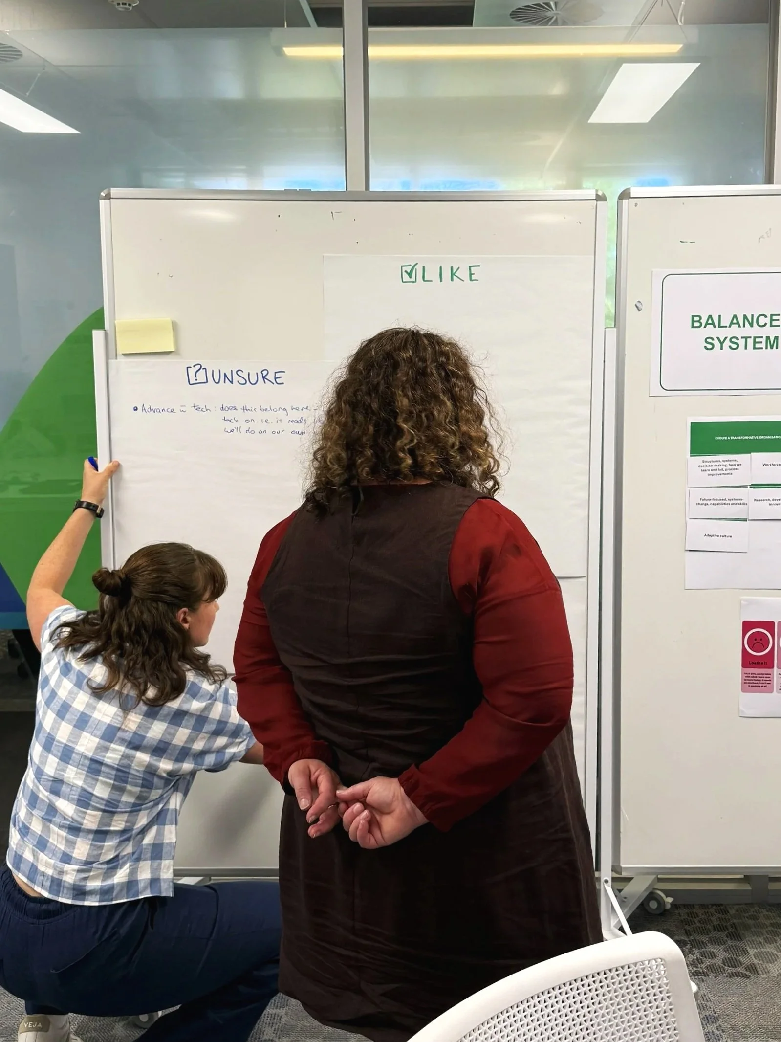 Two people participate in a collaborative workshop activity, using whiteboards labelled ‘Like’ and ‘Unsure’ to capture feedback. One person crouches beside the board to add ideas while another stands nearby.