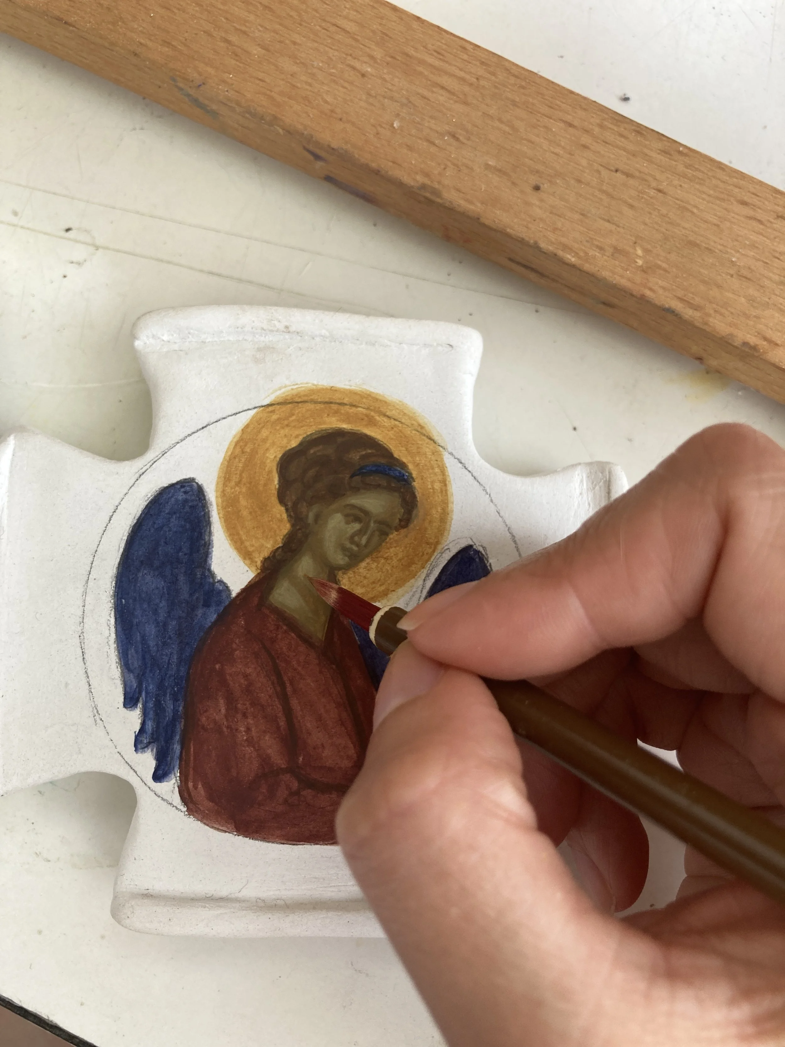 Person painting a religious icon on a-shaped piece of wood, depicting an angel with a golden halo, blue wings, and a red robe.