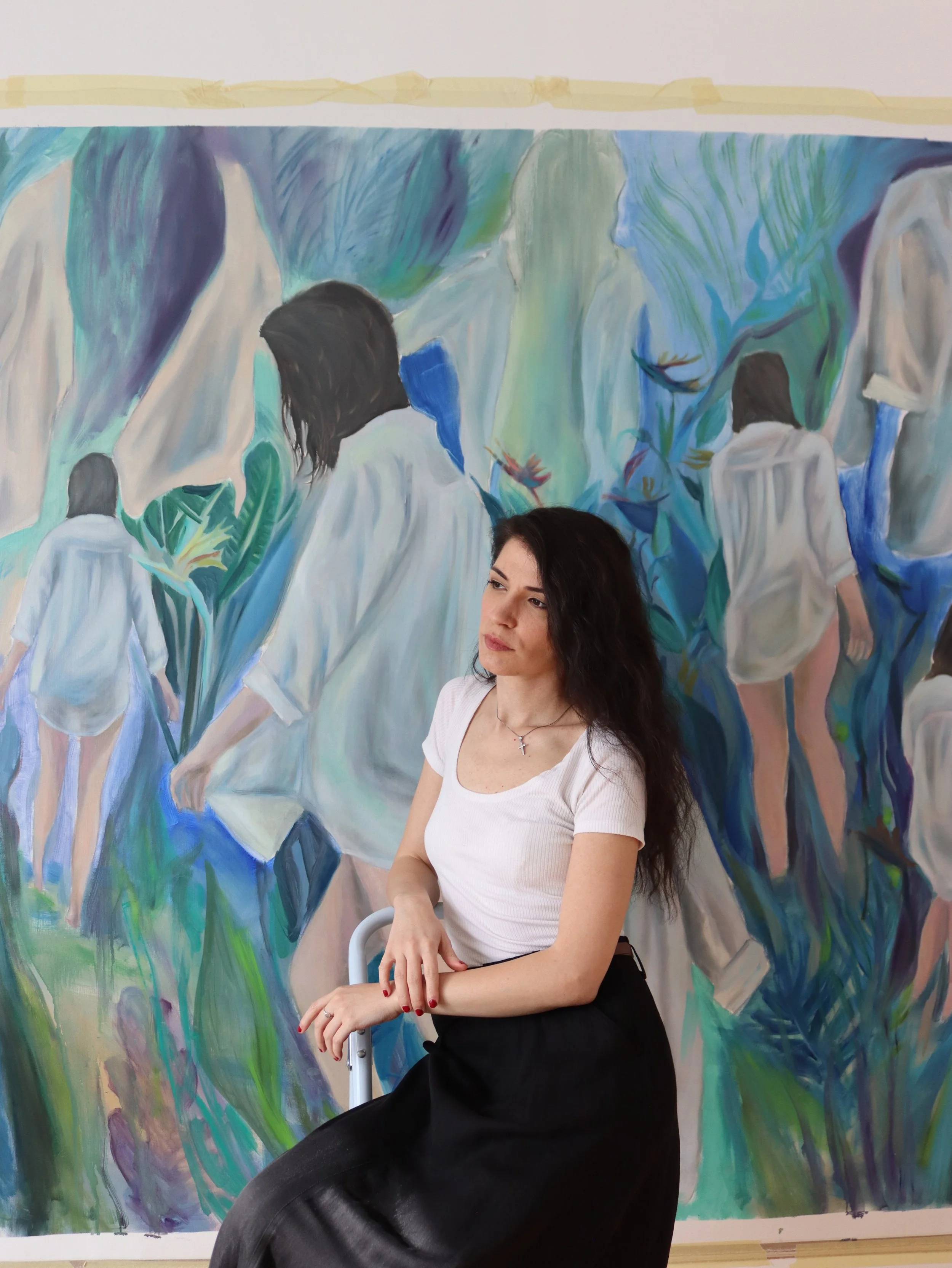 Portrait of the artist, sitting with arms resting on a white cane, in front of a colorful painting with multiple figures and flowing shapes.