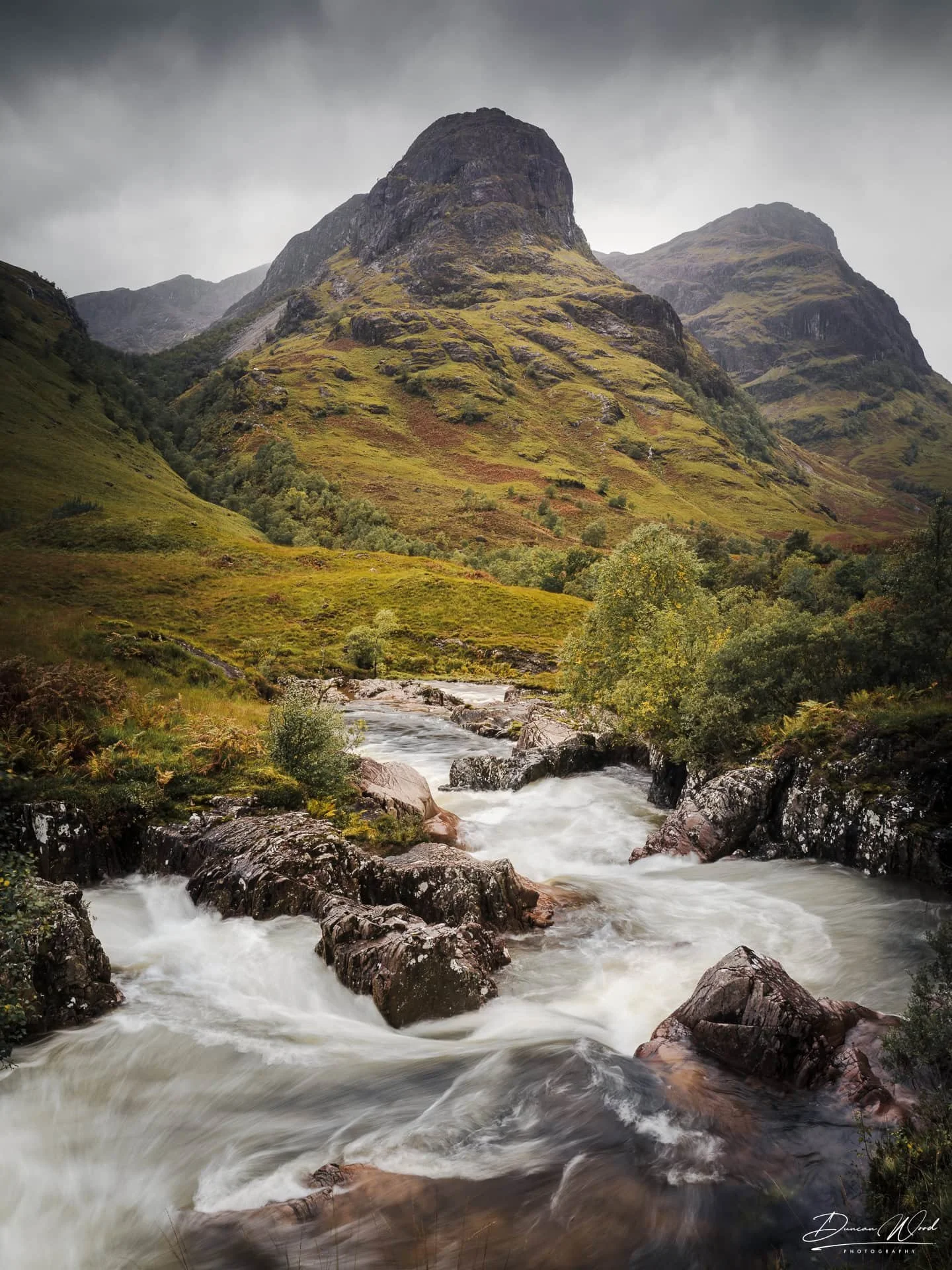 A dramatic view of a rugged mountain peak in Glencoe with a rushing white-water river flowing through a rocky valley in the foreground under a moody sky.