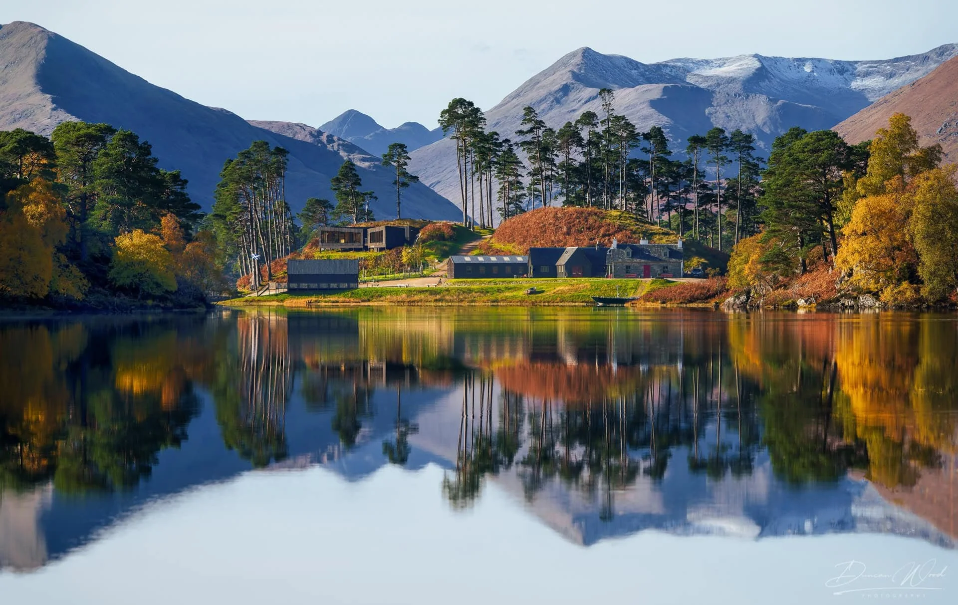 A serene landscape of a lake with autumn-colored trees and a few buildings, surrounded by mountains with some snow on their peaks, under a clear sky.