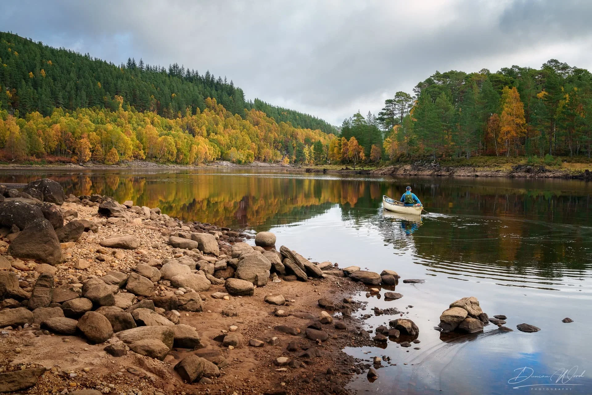 Person in a blue jacket and cap paddling a small white boat on a calm river surrounded by a forest with trees showing fall colors, rocky shoreline in foreground, mountain hills in background, overcast sky.