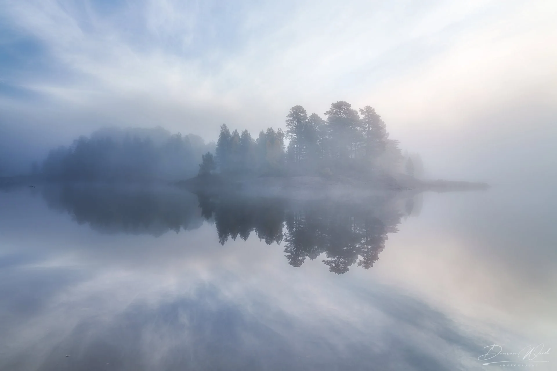 A foggy morning scene of a calm lake with trees and hills reflecting in the water.