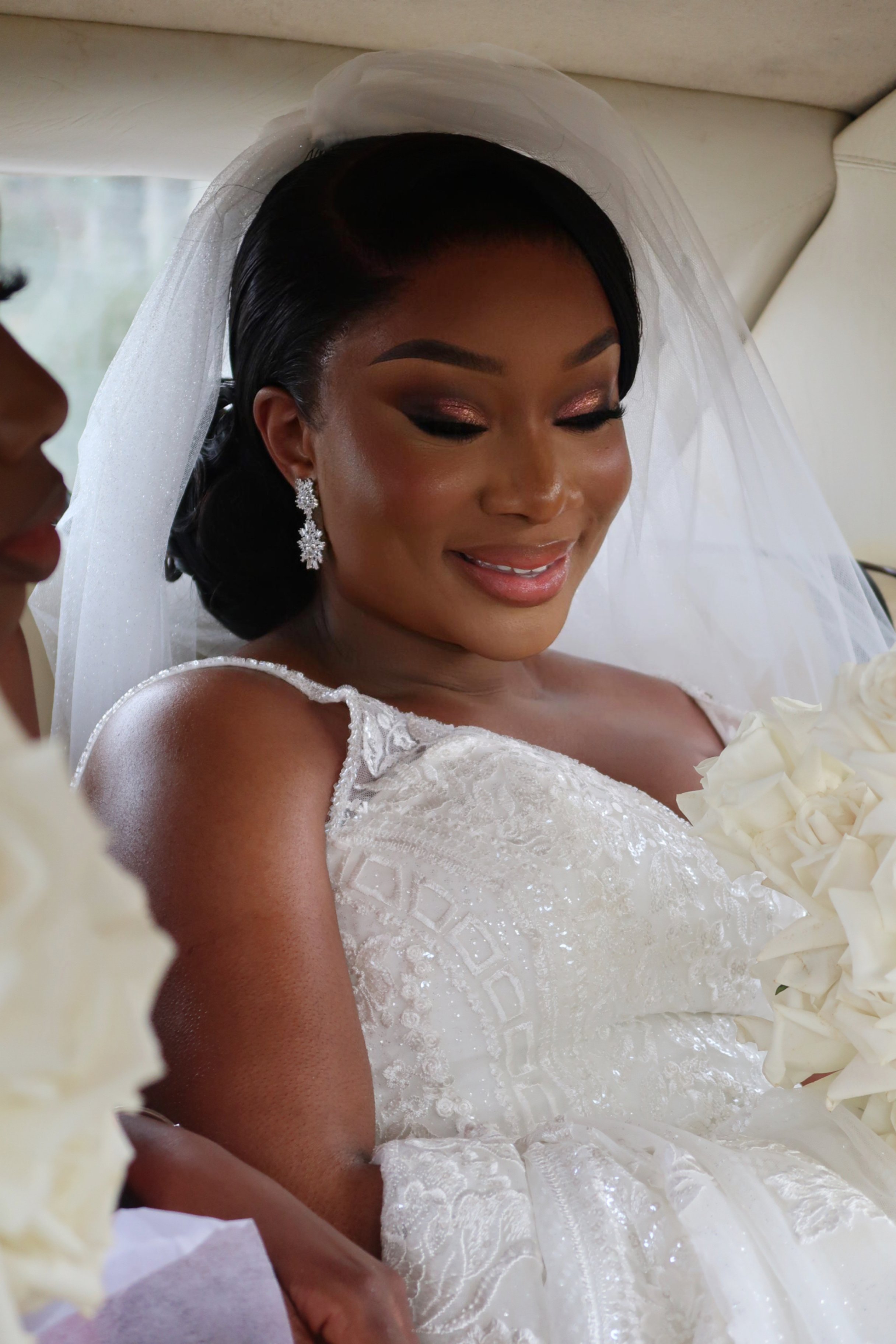 A bride in a white wedding dress with lace details and a veil, holding a bouquet of white flowers, smiling while sitting in a vehicle.