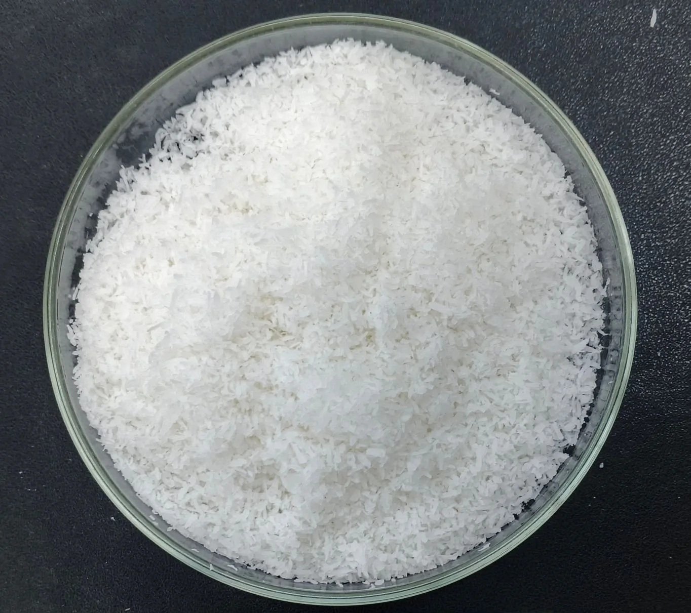 A glass bowl filled with shredded white coconut or desiccated coconuts on a dark surface.