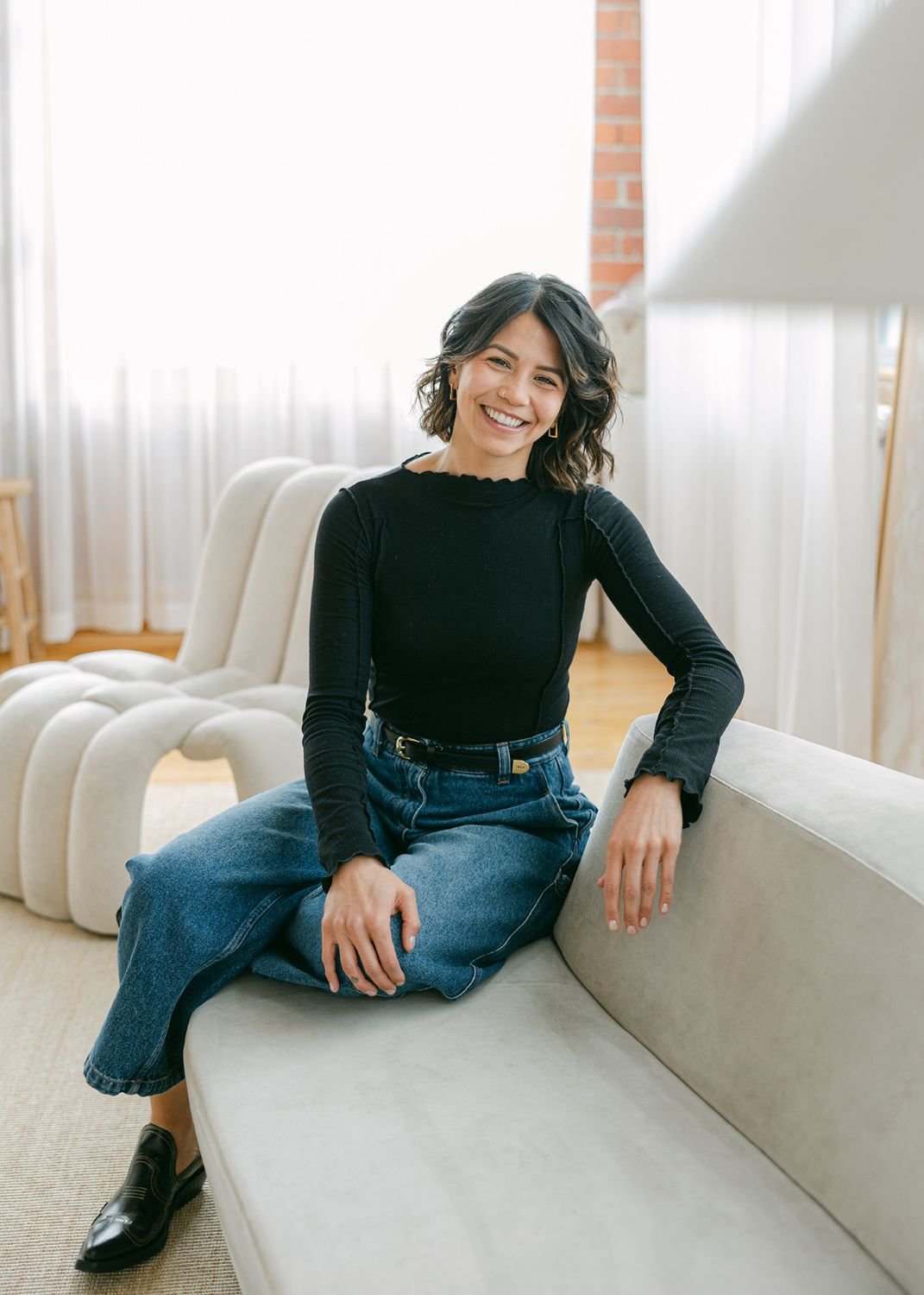 A woman with shoulder-length curly dark hair, wearing a black long-sleeve top, blue jeans, and black shoes, sitting on a beige sofa in a well-lit room with large curtains and a brick wall in the background. She is smiling at the camera.