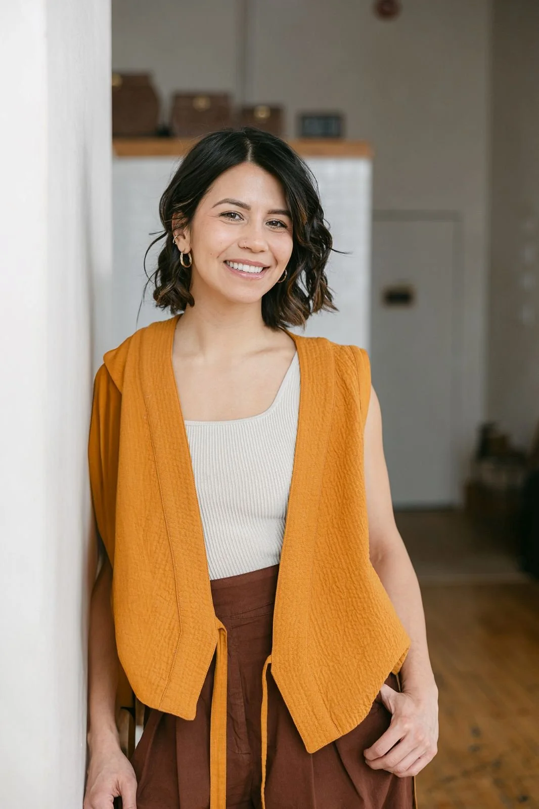 A woman with shoulder-length dark hair, wearing a white top and an orange sleeveless vest, standing indoors and smiling at the camera.