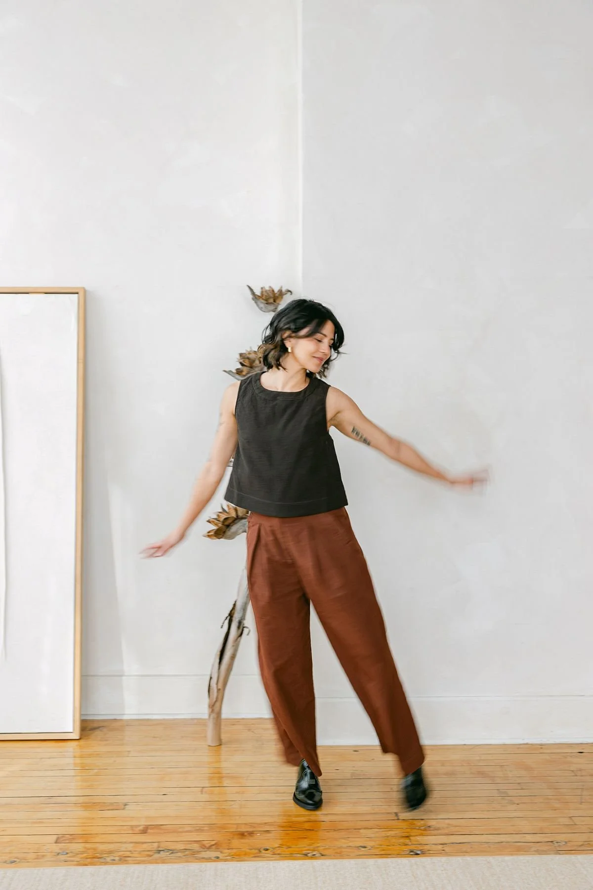 A woman with short black hair, wearing a sleeveless black top, brown pants, and black shoes, is dancing in a minimalistic room with light walls and wooden flooring.