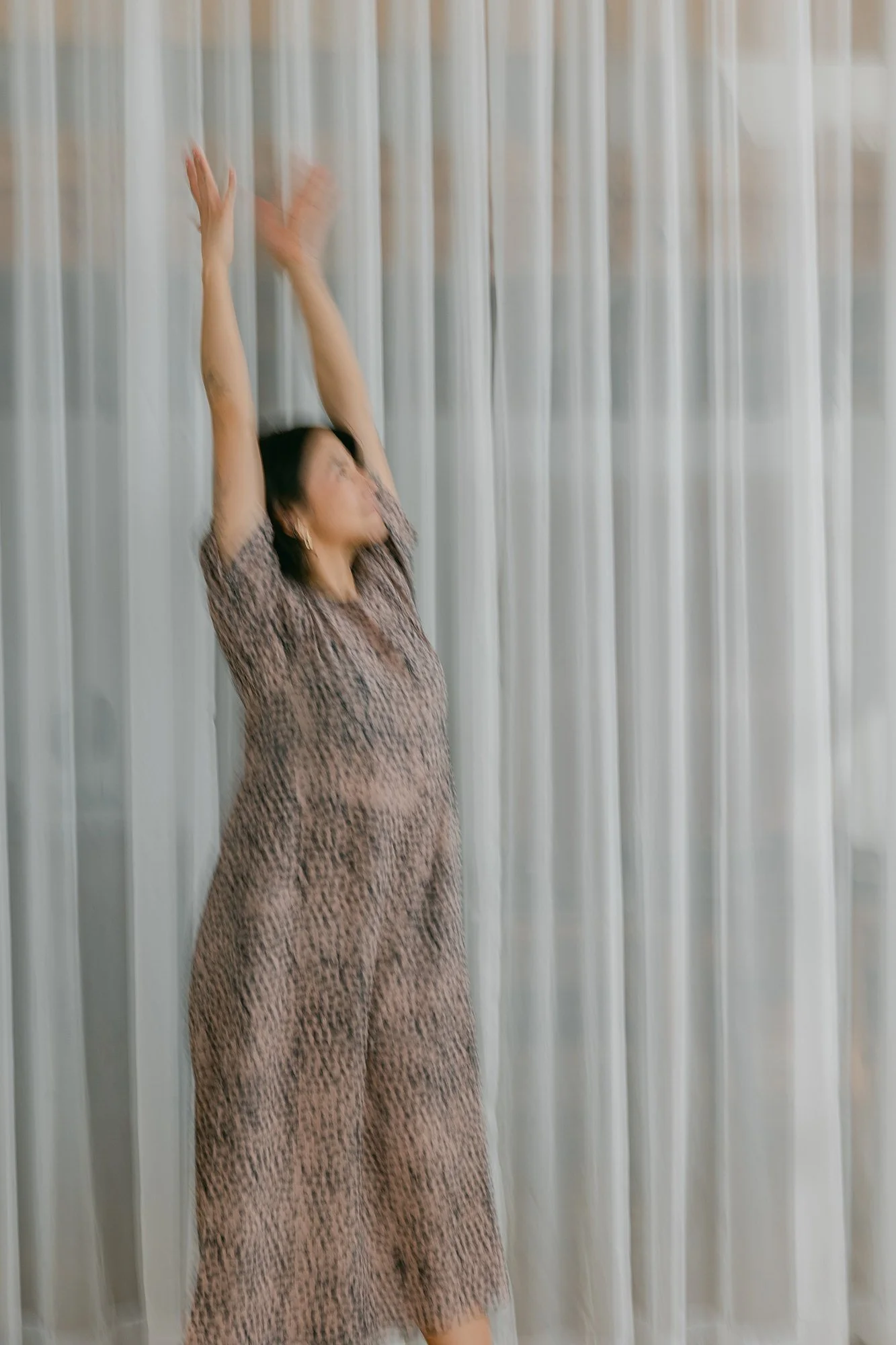 Woman in animal print dress stretching with arms raised in front of a white curtain.