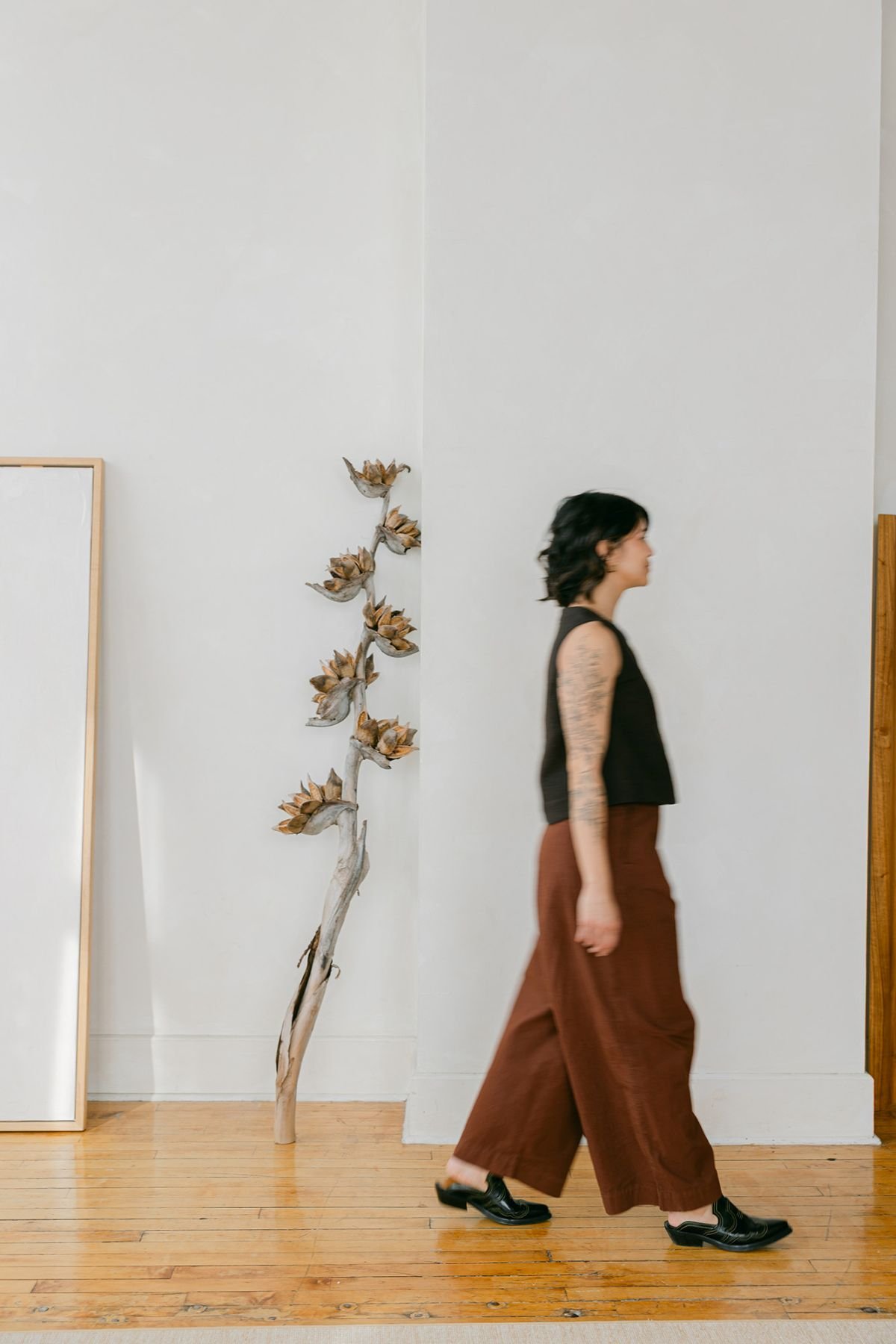A woman with dark curly hair, wearing a sleeveless black top and brown wide-leg pants, walking past a white wall. There is a dried artichoke plant with multiple branches and leaves positioned against the wall, and a partially visible rectangular frame leaning on the wall.