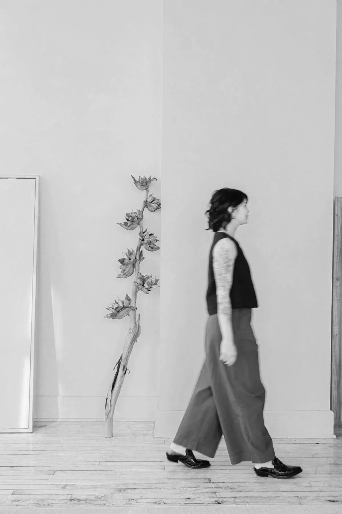 A woman with short wavy hair, wearing a sleeveless black top, wide-leg pants, and black shoes, walking past a painted plant on a wall and a mirror leaning against the wall in a minimalist interior.