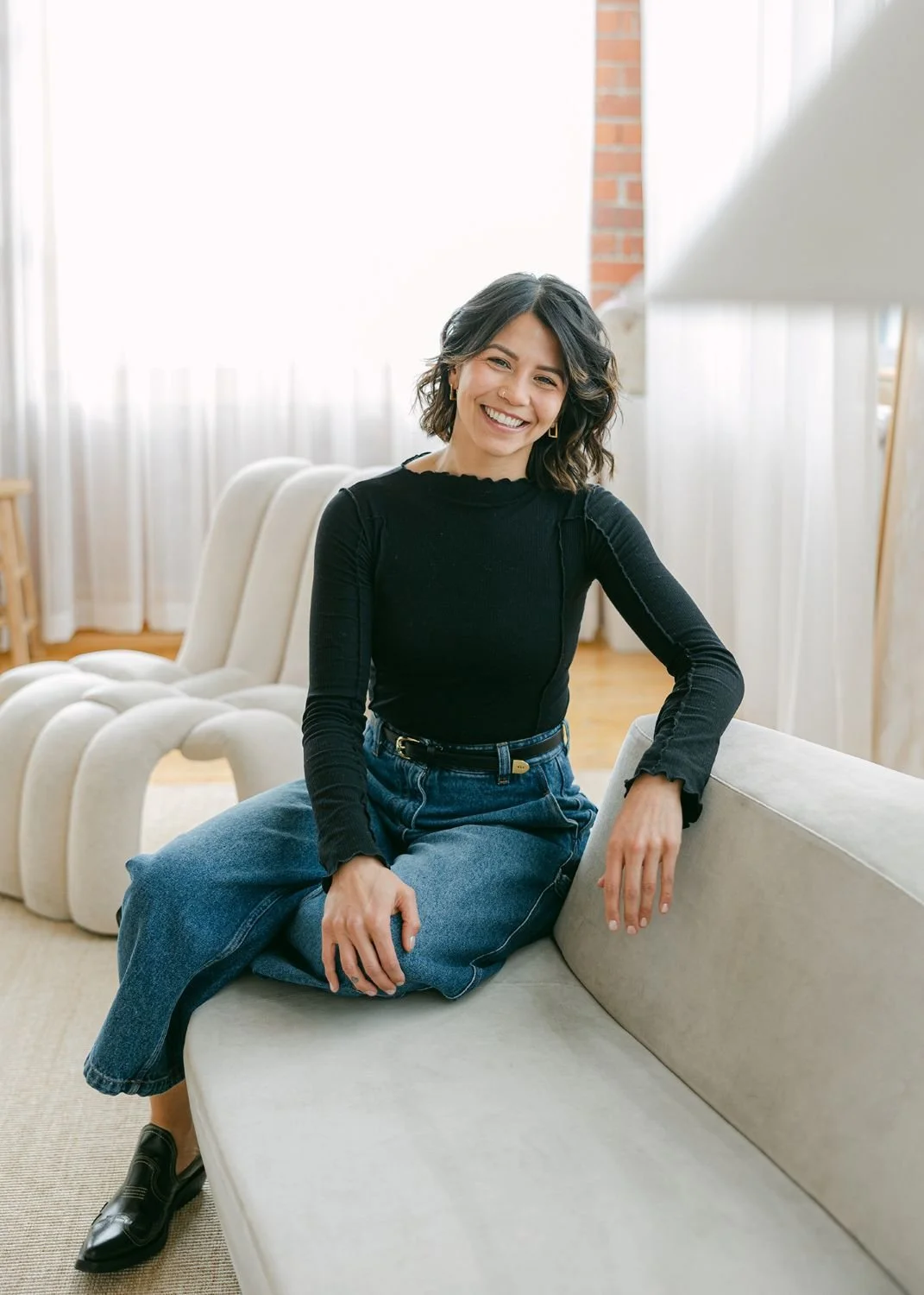 A woman sitting on a sofa in a bright room, smiling, with short wavy dark hair, wearing a black long-sleeve top, blue jeans, and black shoes.