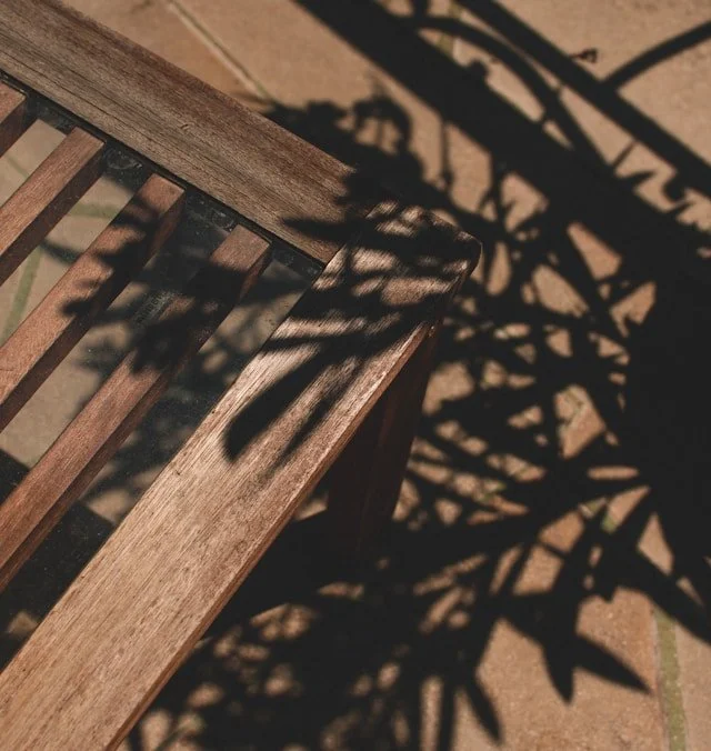 Close-up of a wooden outdoor bench with shadow of nearby plants cast on it, sitting on a brick patio.